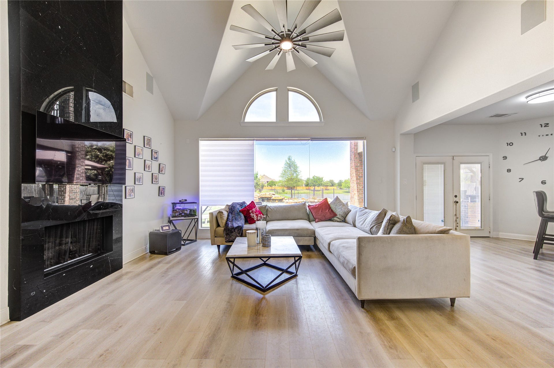 Spacious and inviting living room featuring soaring vaulted ceilings and an abundance of natural light. A dramatic floor-to-ceiling fireplace creates a striking focal point, complemented by modern finishes and warm wood flooring throughout. The oversized windows and sliding glass doors offer beautiful backyard views and seamless indoor-outdoor flow. The open layout provides ample space for comfortable seating and entertaining, while the contemporary light fixture adds a stylish touch to the airy atmosphere.