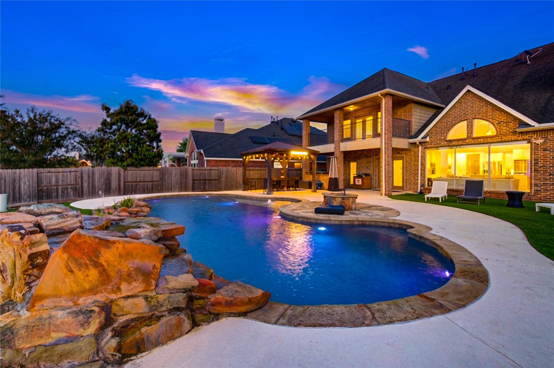 Resort-style backyard at twilight featuring a stunning lagoon-style pool with rock waterfall, ambient lighting, and beautifully landscaped surroundings. The illuminated home and outdoor living spaces create a perfect setting for evening entertaining with a luxurious, private ambiance.