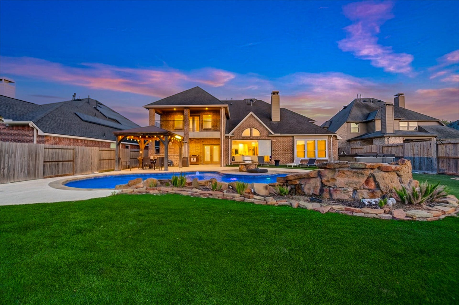 Stunning backyard oasis designed for luxury living and unforgettable entertaining. This beautifully landscaped space features a sparkling resort-style pool with water features, a spacious patio, and a fully equipped outdoor kitchen. The covered pavilion offers a perfect spot to relax or host gatherings, complete with warm lighting and elegant finishes. Expansive green space provides room to play or unwind, all set against a breathtaking sunset backdrop that creates a truly serene and private retreat.