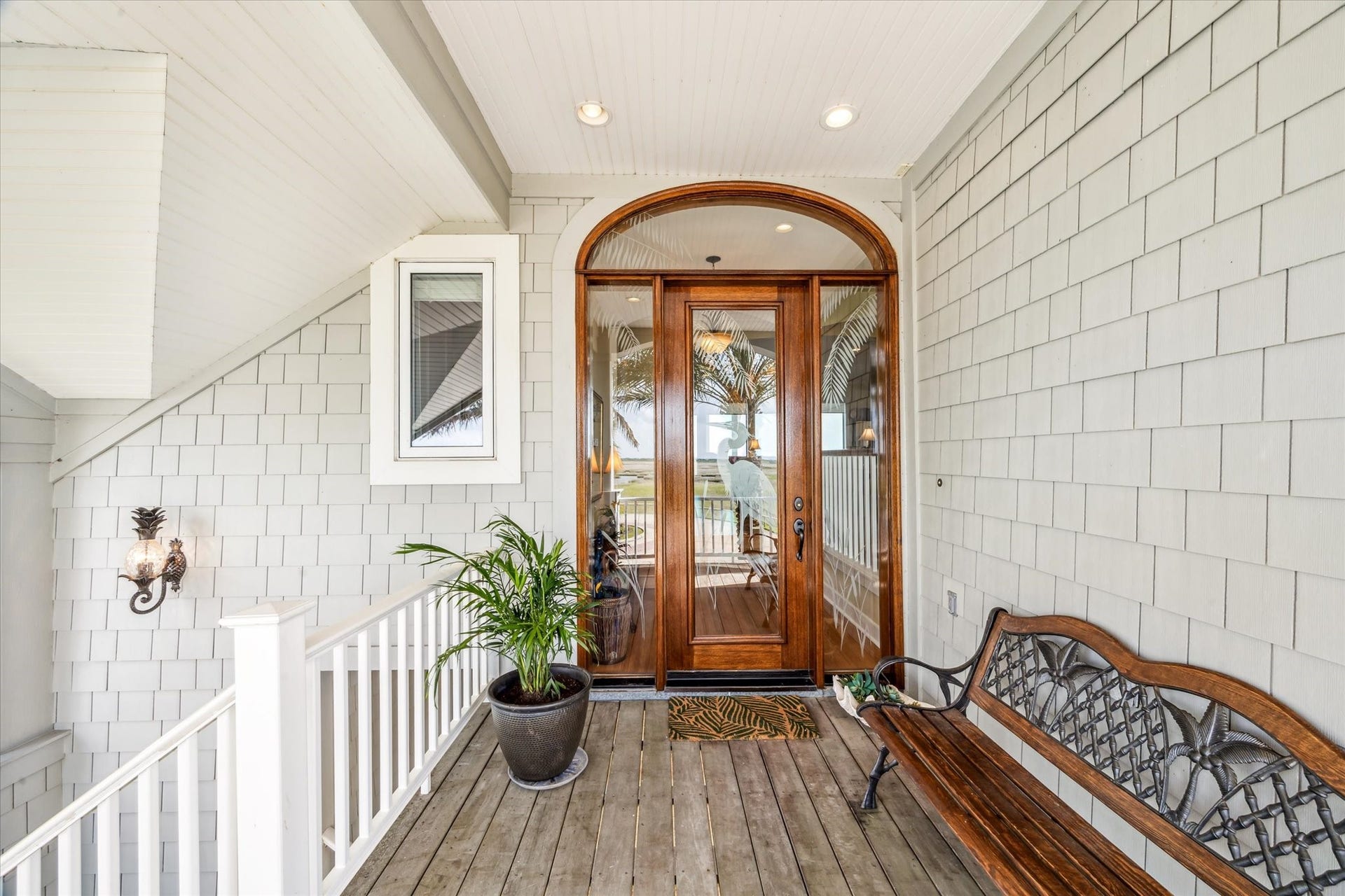 1 Shell Key, Hitchcock, TX 77563 Welcome guests in style with this inviting front entry, featuring rich wood-framed etched glass door beneath an elegant arched transom. The covered porch is accented with low-maintenance Hardie shingle siding, warm-toned IPE decking, and coastal-inspired details including a decorative wall sconce and classic white railing, making this space the perfect transition into the relaxed yet refined interior of the home