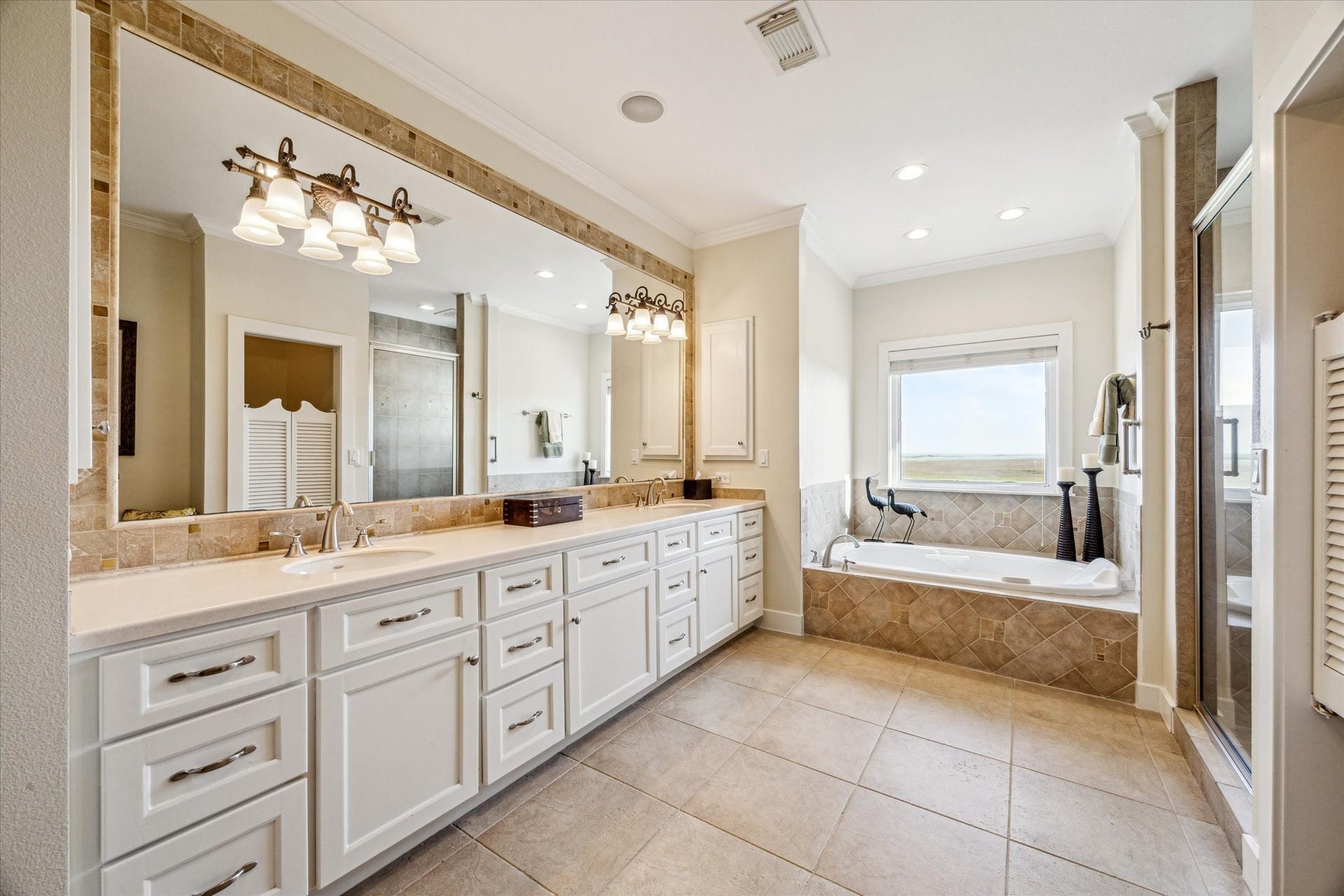 1 Shell Key, Hitchcock, TX 77563 This elegant bathroom features a spacious, spa-like design with a large dual vanity topped with sleek countertops and framed by a full-width mirror with decorative lighting. The custom cabinetry offers abundant storage with a clean, white finish that complements the neutral tile flooring.
