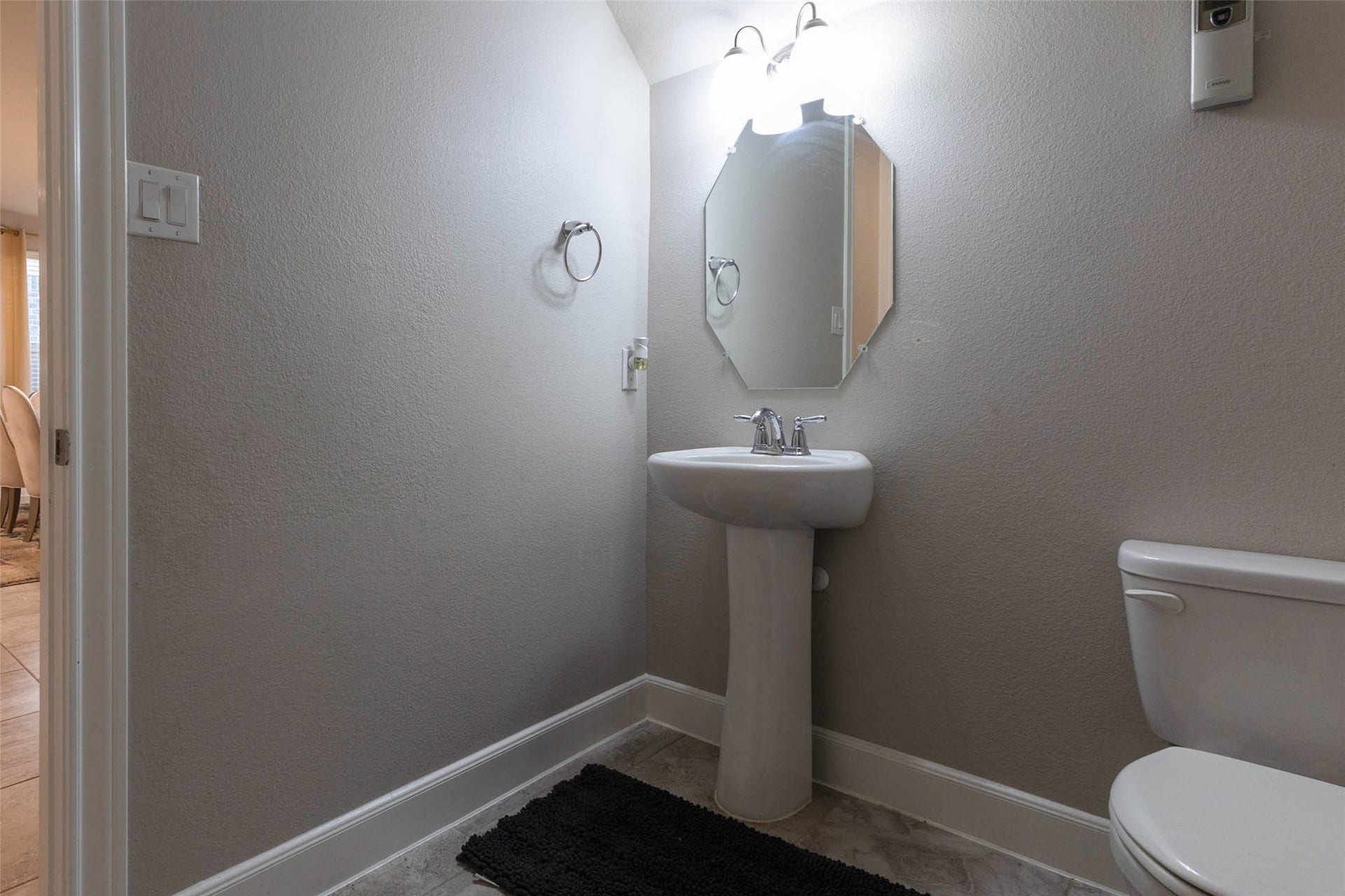 A guest powder bath with a pedestal sink, an octagonal framed mirror and a toilet in a compact, convenient layout. Warm gray walls and wall sconces give the room a finished look, and the doorway on the left leads back to the living space. A powder bath like this keeps guests out of the private bedrooms and bathrooms. It is small, tidy and ready for entertaining. Perfect for guests during a party or family gathering.