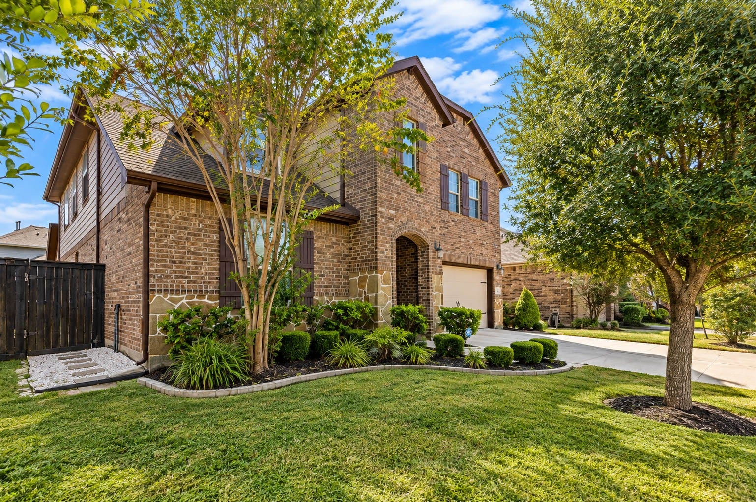 The front corner view captures the brick two-story facade, the arched front entry and a driveway framed by crape myrtles and manicured landscaping beds. Healthy grass and mature trees give the street-side presence a settled, established feel. Built in 2019 in Harvest Green, this home welcomes you with warm brick tones, dark shutters and a garage that sits neatly to one side of the front elevation.