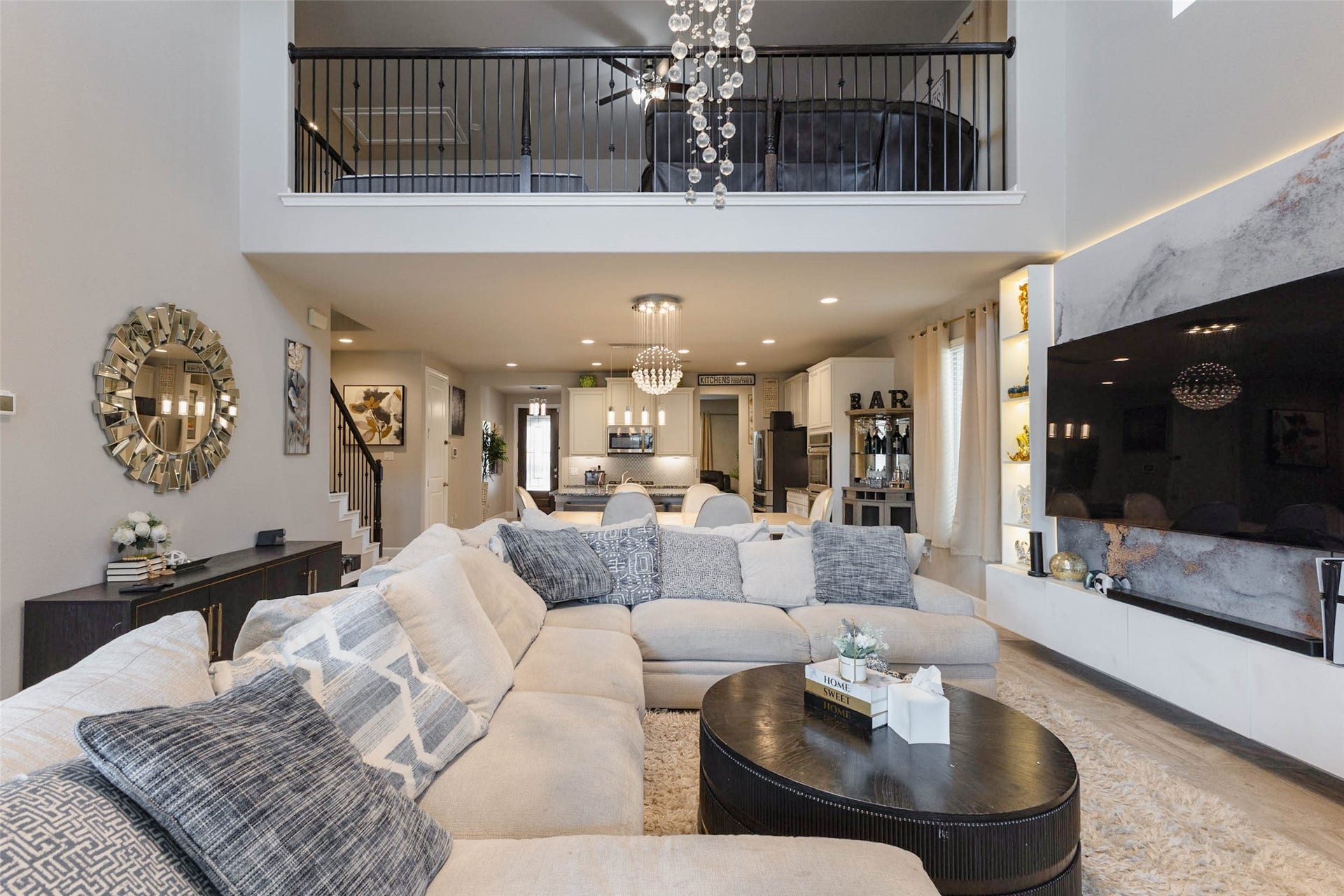 A wider look at the family room shows the built-in media wall with accent lighting and shelving, plenty of floor space for a large sectional and a clear view back to the kitchen and dining area. The upstairs railing runs along the open ceiling, adding architectural interest. Tile flooring keeps the main living area durable and easy to clean. It is a room made for game days, movie nights and holiday gatherings.