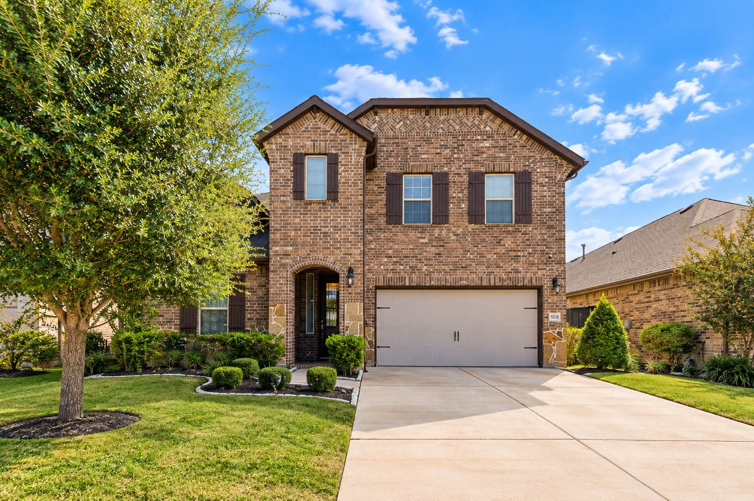 Straight-on front elevation with a covered arched entry, dark shutters on the upstairs windows and a wide driveway leading to the garage. Brick covers the full facade, which keeps maintenance low and gives the house a classic look. Landscaping beds line the walkway and a mature tree adds shade on the left. The proportions are balanced and welcoming, and the clear blue Texas sky makes the colors pop.