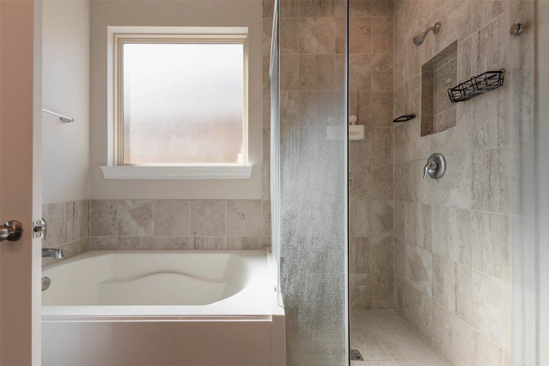 A soaking tub sits beside a frosted window and a glass-enclosed shower with a tiled surround and a built-in niche. Warm, natural-toned tile covers the shower walls and the tub deck, which gives the room a spa-like quality. The separate tub and shower let you choose between a long soak and a quick rinse. It is a peaceful, private space at the end of the day.