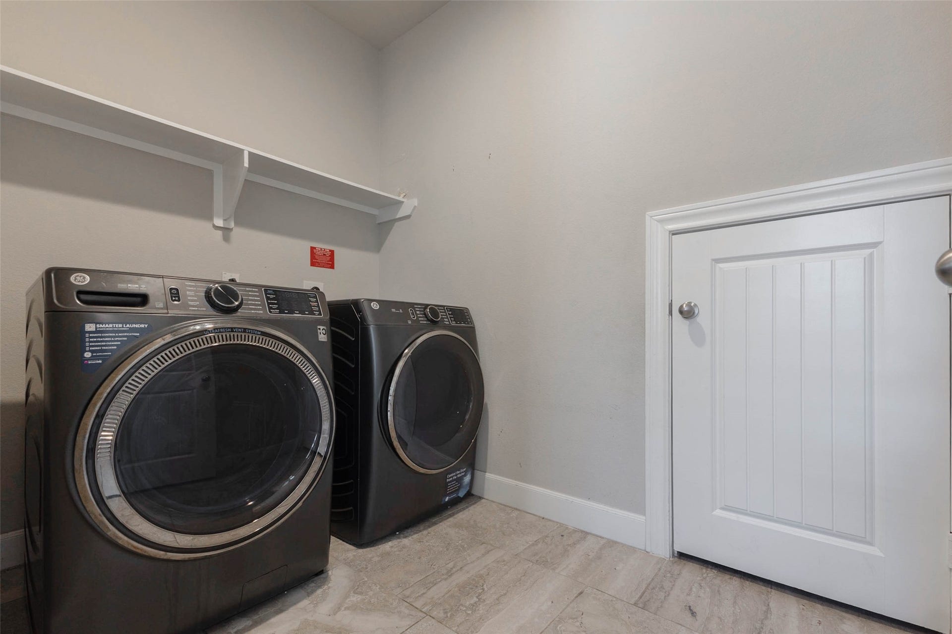 The laundry room has tile flooring, a shelf and rod above the machines and a door to the hallway. There is room for a full-size front-load washer and dryer side by side, plus space nearby for a hamper or folding. A dedicated laundry room keeps this household chore simple and tidy, and the shelf above is handy for detergent and supplies.