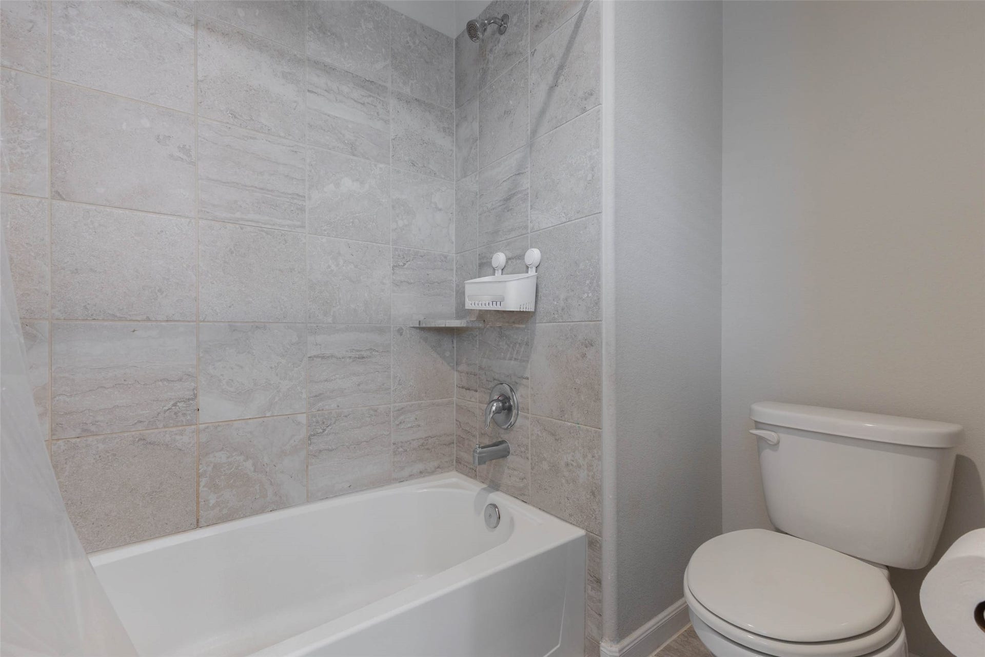 A tub and shower combination with a tiled surround, a soap shelf and a toilet beside it. Large gray tiles run floor to ceiling around the tub, which makes cleaning easy and gives the room a modern feel. A window lets in light, and the bath is conveniently located near the bedrooms. This is a practical bathroom for kids, guests or anyone who prefers a classic bath and shower setup. Easy to keep clean and a comfortable fit for the whole family.