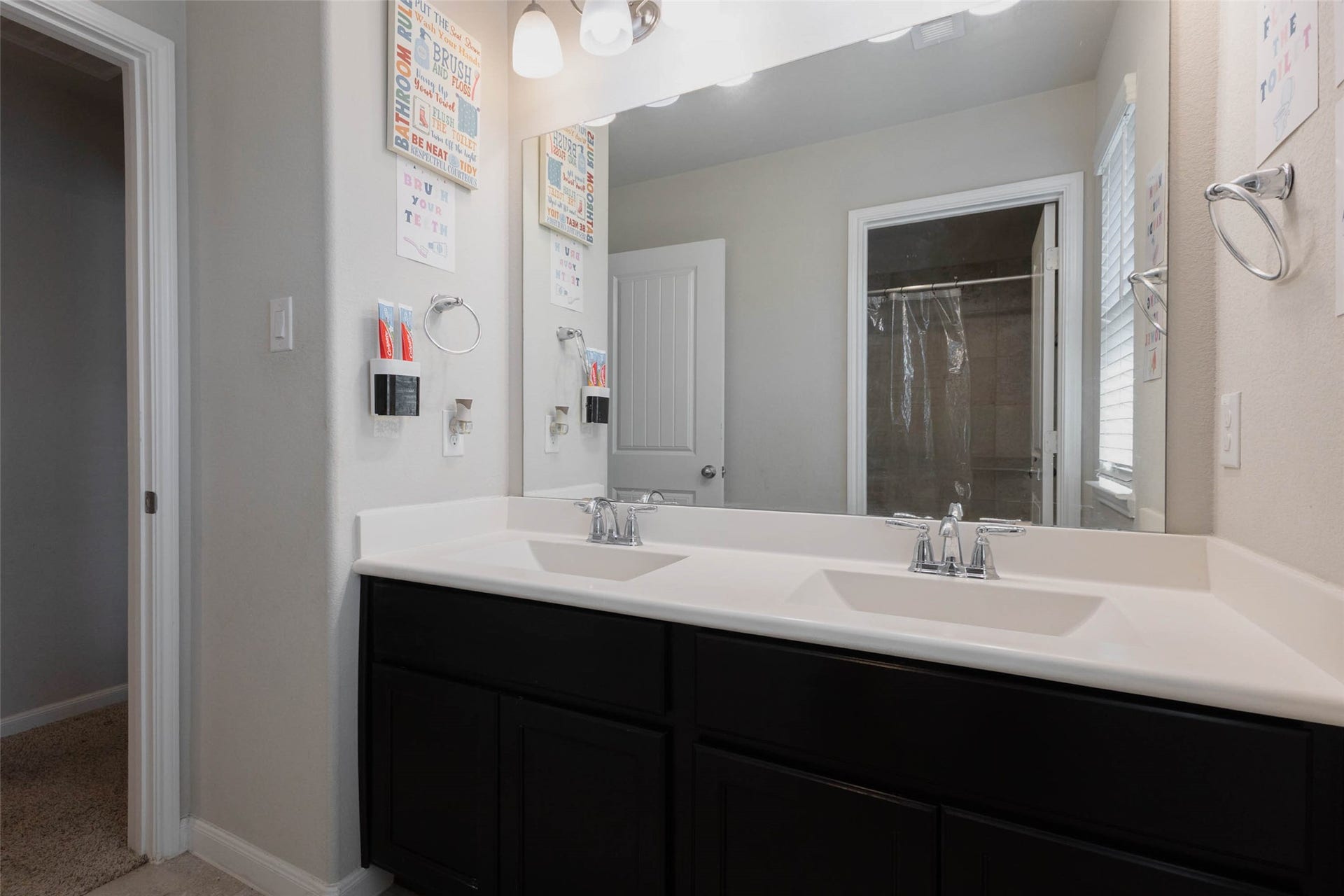 A bathroom with a long dual-sink vanity, dark cabinetry and a white countertop, plus a large mirror with a lighted fixture above. A shower enclosure sits behind the mirror and a doorway opens to the hall. Two sinks help busy mornings run smoothly, and the dark cabinets provide plenty of storage underneath. The contrast between dark and light finishes gives the space a modern look. Everything is within easy reach in this well-planned space.