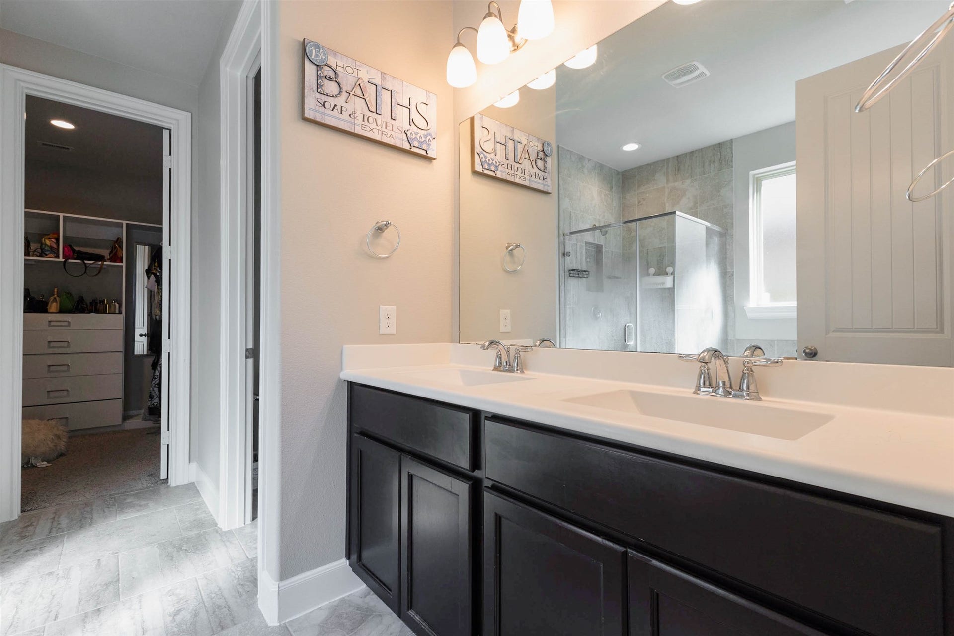 A bathroom with a long double vanity in dark cabinetry, a white countertop, wall sconces and a large mirror. A doorway on the left leads to a walk-in closet with built-in shelving visible in the background, and a glass shower enclosure sits at the end of the room. Dual sinks give each person their own space. This is a comfortable and well-organized bathroom for the primary suite. It adds a real sense of luxury to the daily routine.