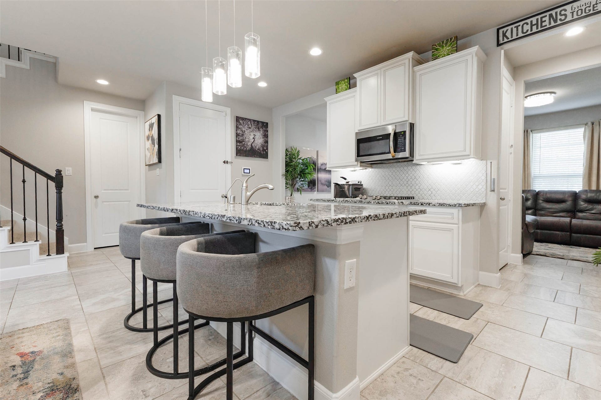 The kitchen island has a granite top, three bar stools and pendant lights hanging above, perfect for casual meals and conversation. White cabinets, a stainless microwave and under-cabinet lighting give the room a bright, clean look. A pantry door and a staircase sit near the kitchen, which keeps storage close and traffic flowing. The seating at the island lets guests visit with the cook without crowding the work zone.