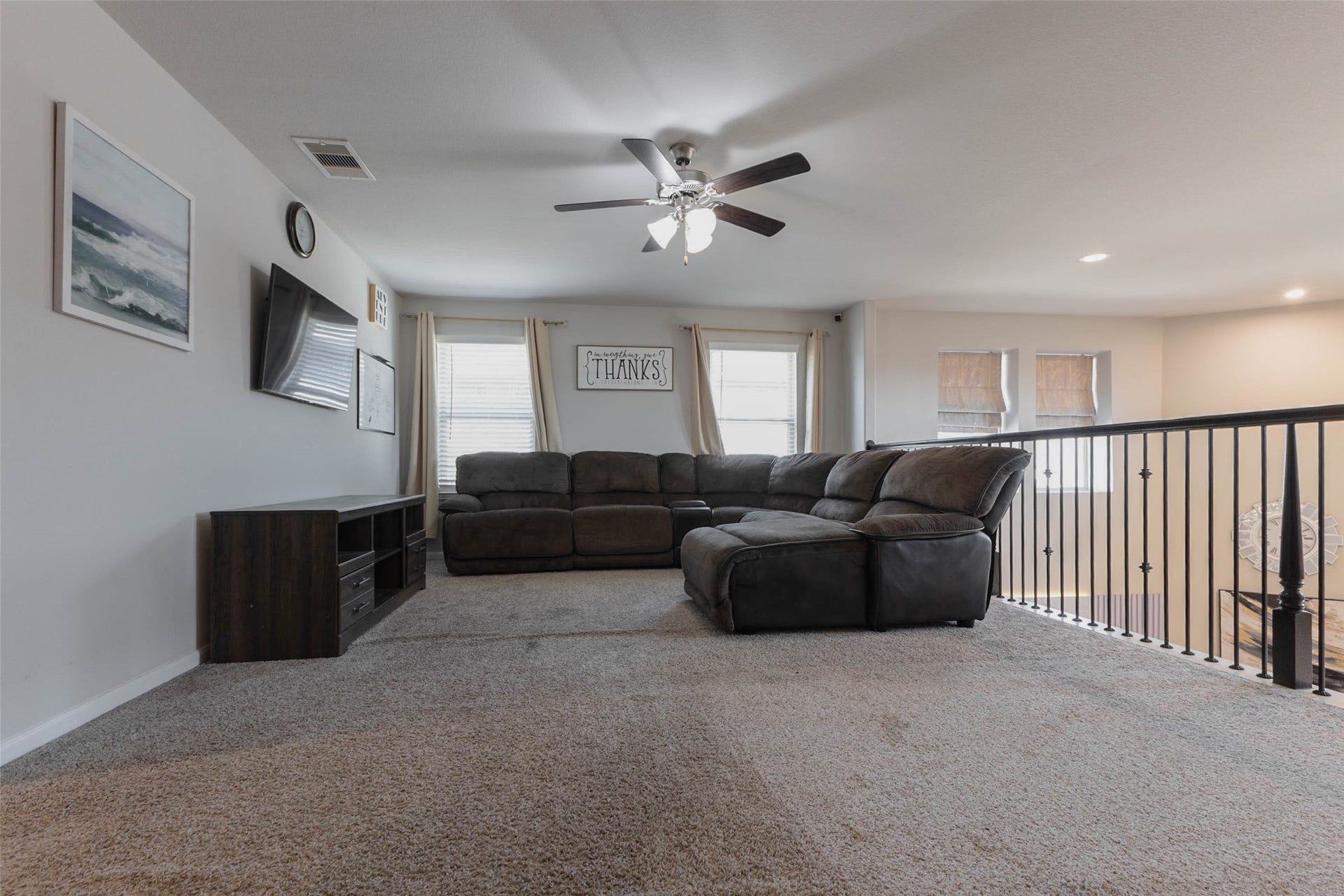 The upstairs game room has a wide open floor plan with an iron railing overlooking the living area below. Large windows bring in daylight, a ceiling fan keeps the air moving and there is plenty of space for a sectional, a game table or a media setup. Carpet makes the room cozy for movie nights and play. A loft-style room like this gives the family a place to gather that feels separate from downstairs.