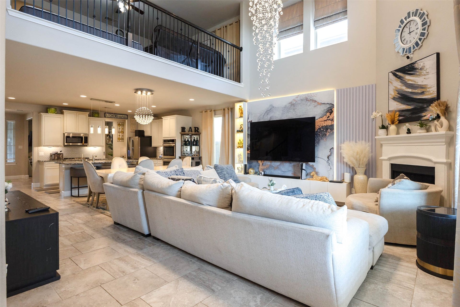 The living room rises two stories, with a catwalk railing on the second floor overlooking the space and a stunning cascading light fixture above. A white sectional anchors the room beside a fireplace with a white mantel, and tall windows pour daylight across the tile floors. The kitchen opens beyond, so the cook stays part of the gathering. This is the room that makes a first impression at every showing.