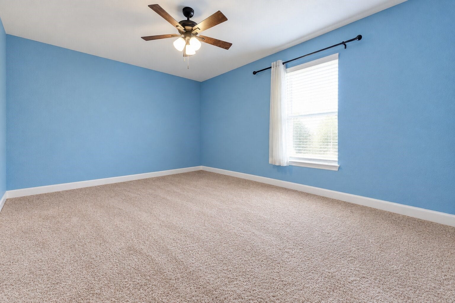 Cheerful bedroom featuring coral-colored walls, soft carpeting, a ceiling fan with lighting, and a window with blinds and curtains. The open layout offers flexibility for a bedroom, creative studio, or home office. Photo has been digitally decluttered.