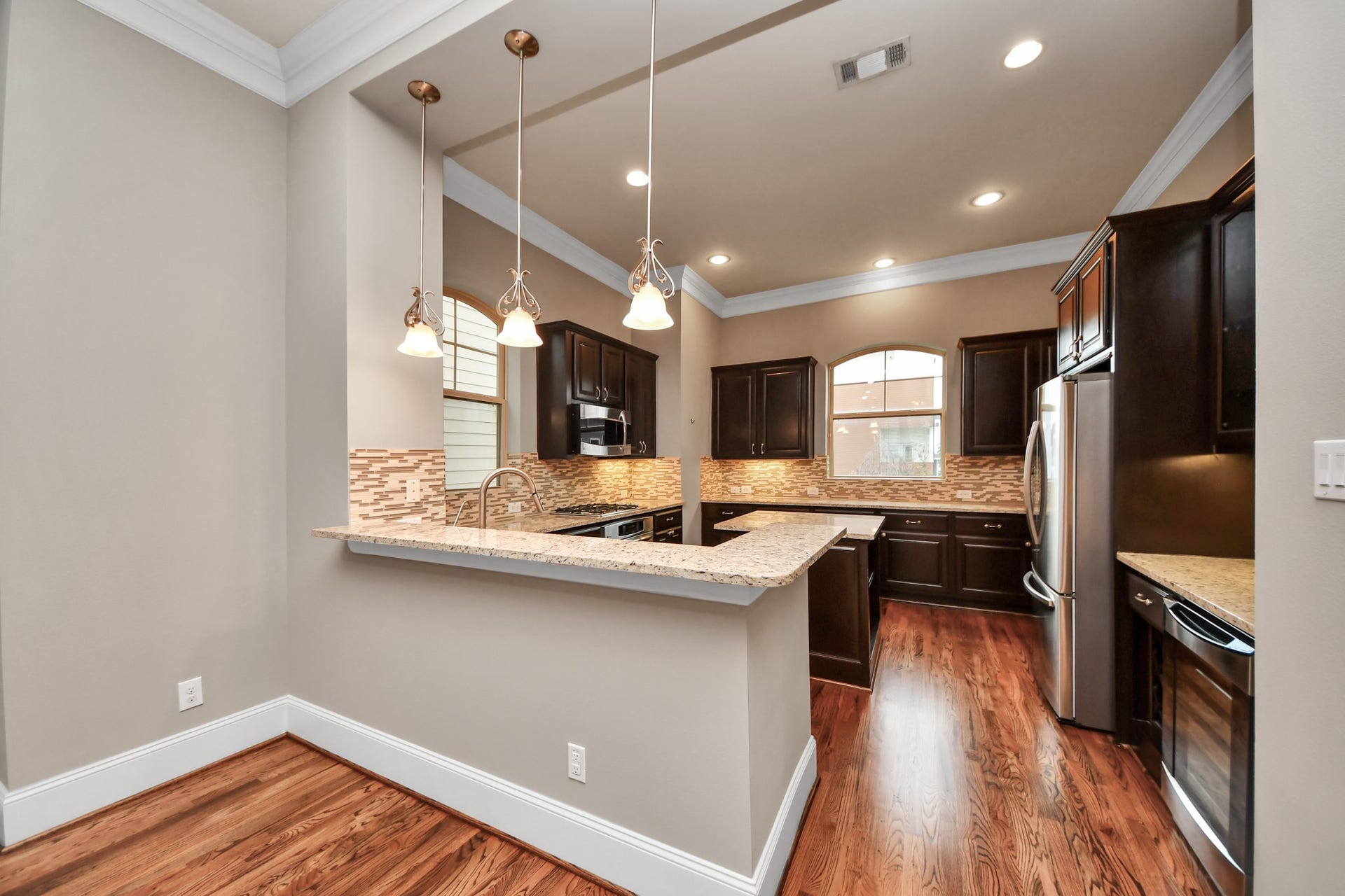 1617 W 23rd Street, Houston, TX 77008 Elegant kitchen featuring rich wood cabinetry, granite countertops, and modern stainless steel appliances. The open layout includes a breakfast bar with pendant lighting, complemented by warm hardwood floors and ample natural light from arched windows.