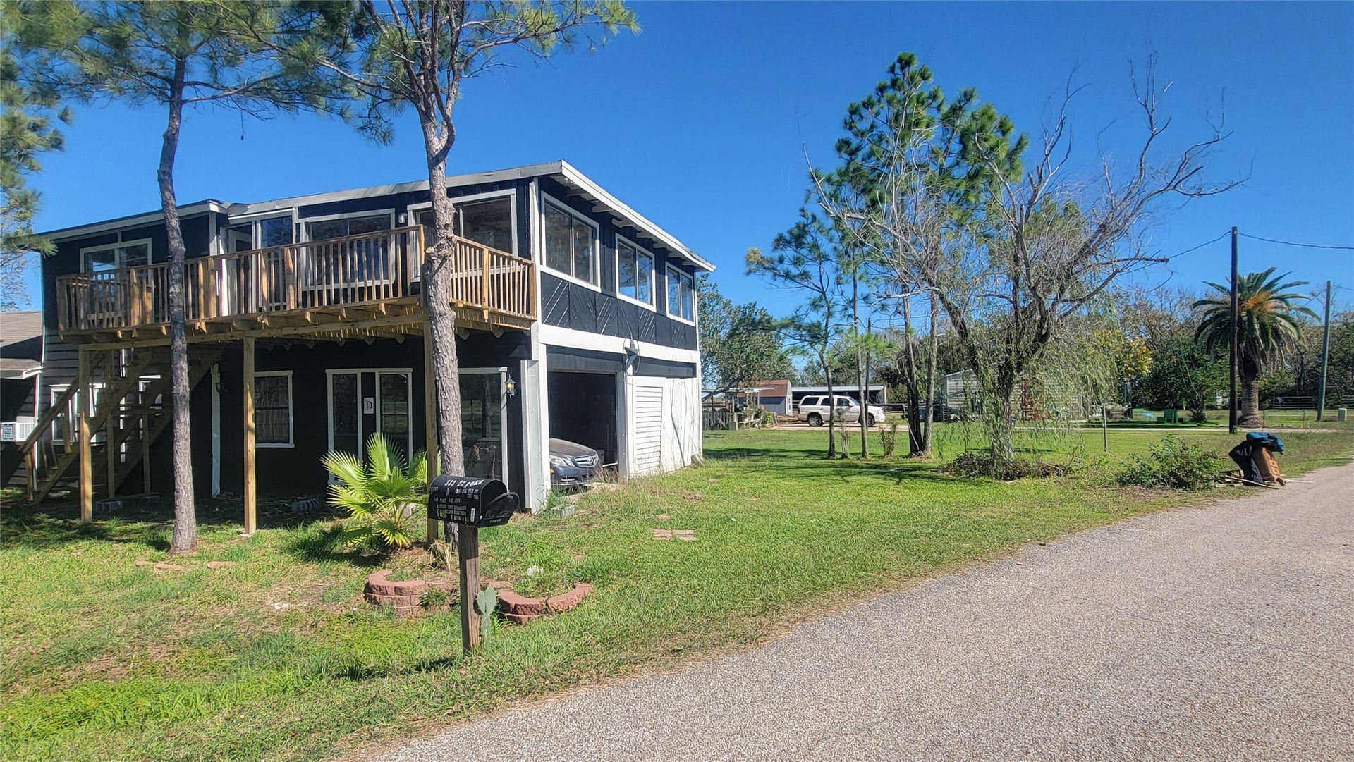 12018 Sandpiper Road, Angleton, TX 77515 This photo shows a two-story home with a wooden deck and enclosed porch on the upper level. It's surrounded by a grassy yard with trees, positioned on a quiet street. The exterior is a mix of dark and light colors, offering a rustic charm.