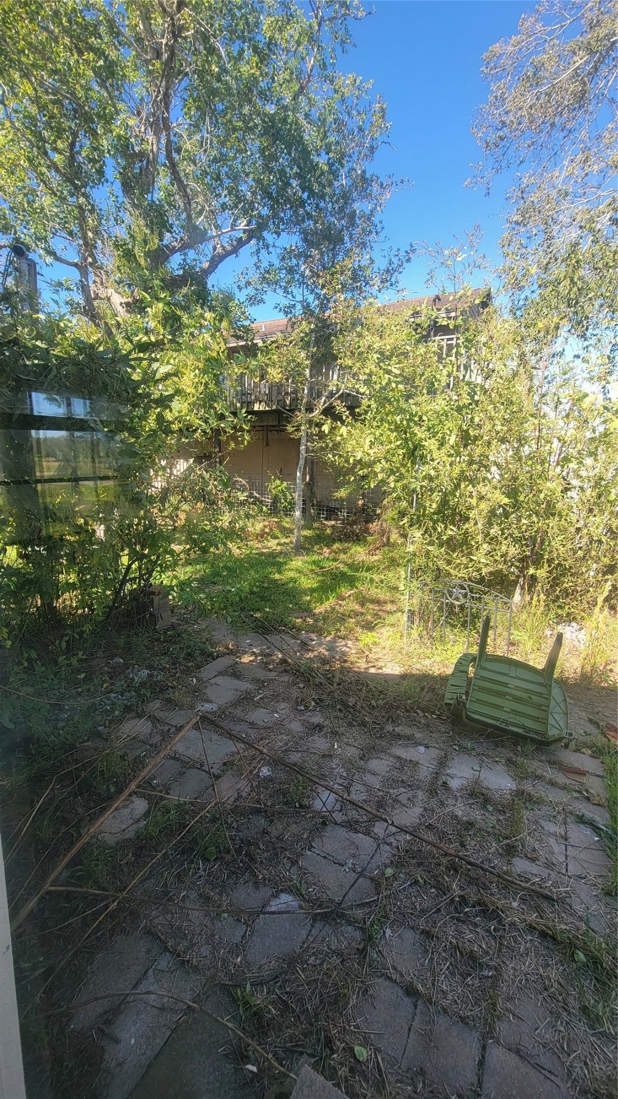 12018 Sandpiper Road, Angleton, TX 77515 This photo shows a backyard area with a stone patio and overgrown vegetation. There are trees providing shade and a view of a neighboring building. The space has potential for landscaping and outdoor activities.