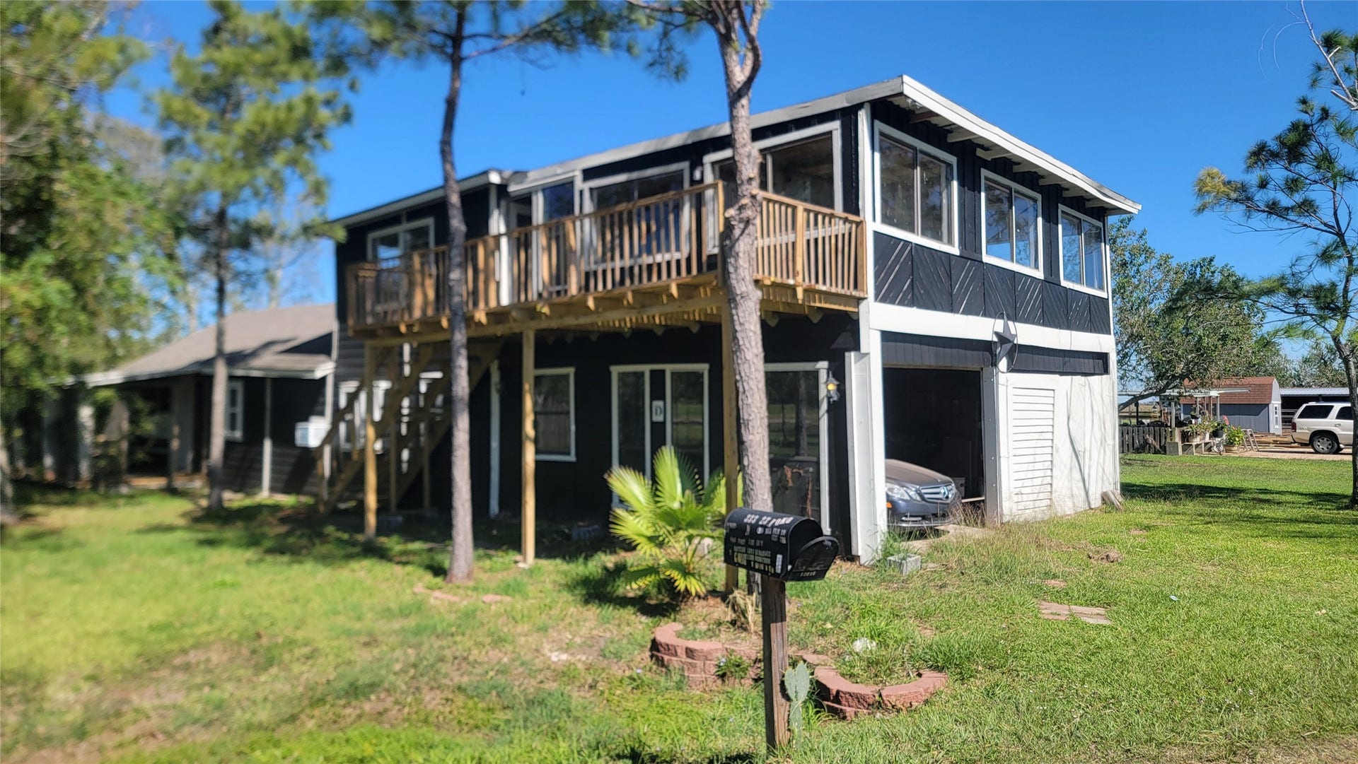 12018 Sandpiper Road, Angleton, TX 77515 This photo shows a two-story home with a dark exterior and white trim. It features an upper-level balcony with large windows, a garage, and is surrounded by a grassy yard with trees.