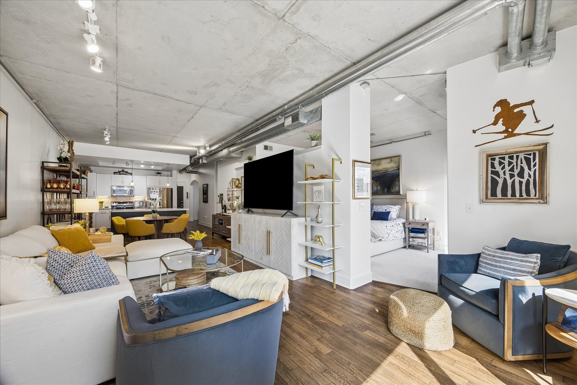 Living area with exposed concrete ceilings, sleek hardwood floors, and abundant natural light. The space seamlessly flows into a contemporary kitchen and cozy bedroom nook, creating an urban retreat.