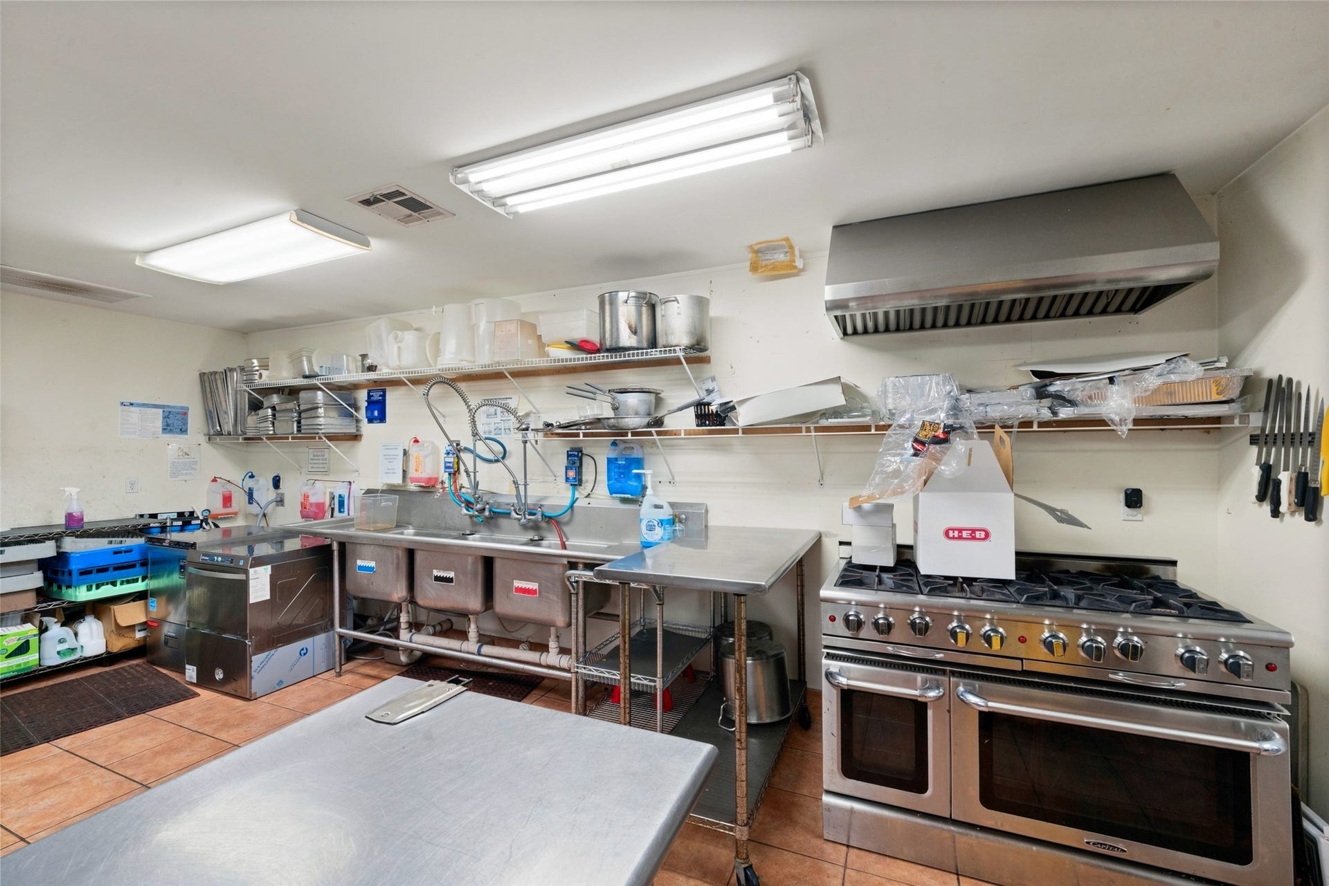 Reception hall kitchen featuring a range beneath a vent hood, stainless steel work surfaces, and extensive shelving for preparation and storage.
