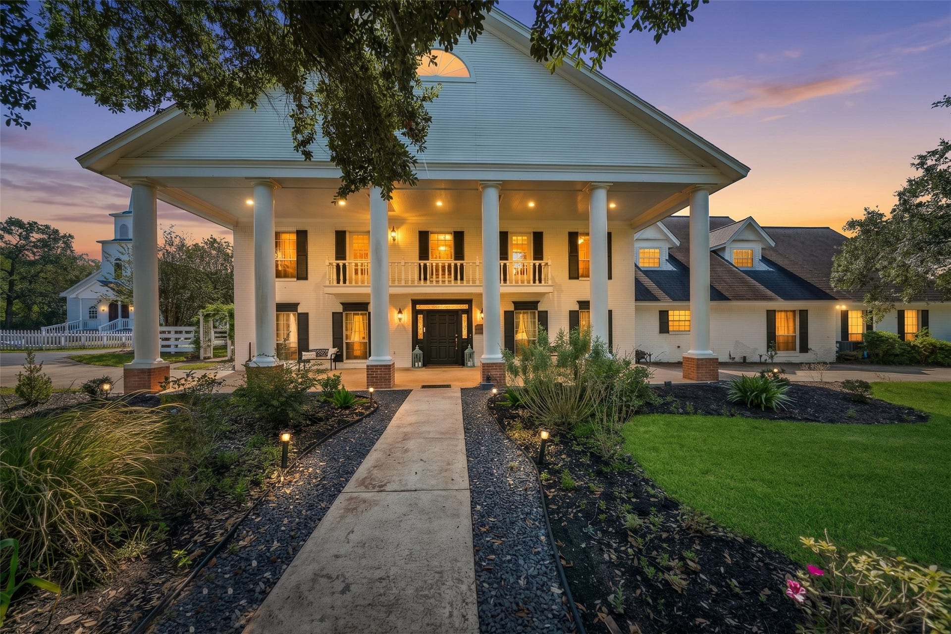 Twilight view of the stately manor, showcasing tall white columns, inviting exterior lighting, and a landscaped walkway.