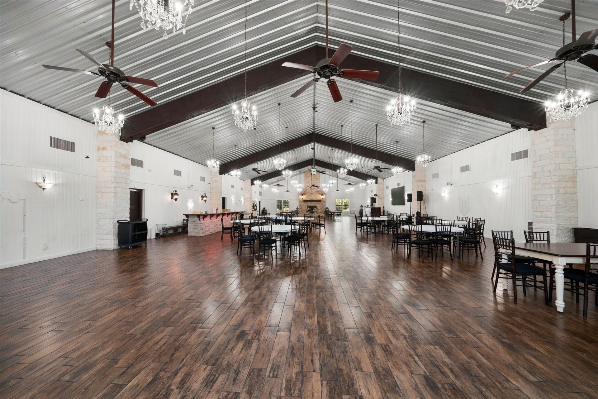 Spacious reception hall featuring a vaulted ceiling, exposed beams, chandeliers, ceiling fans, and an open layout for tables and dancing.
