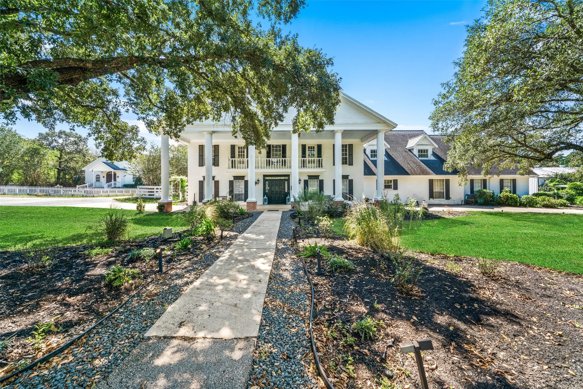 Grand two-story manor framed by mature trees, with a columned entrance and stone walkway leading to the front doors.