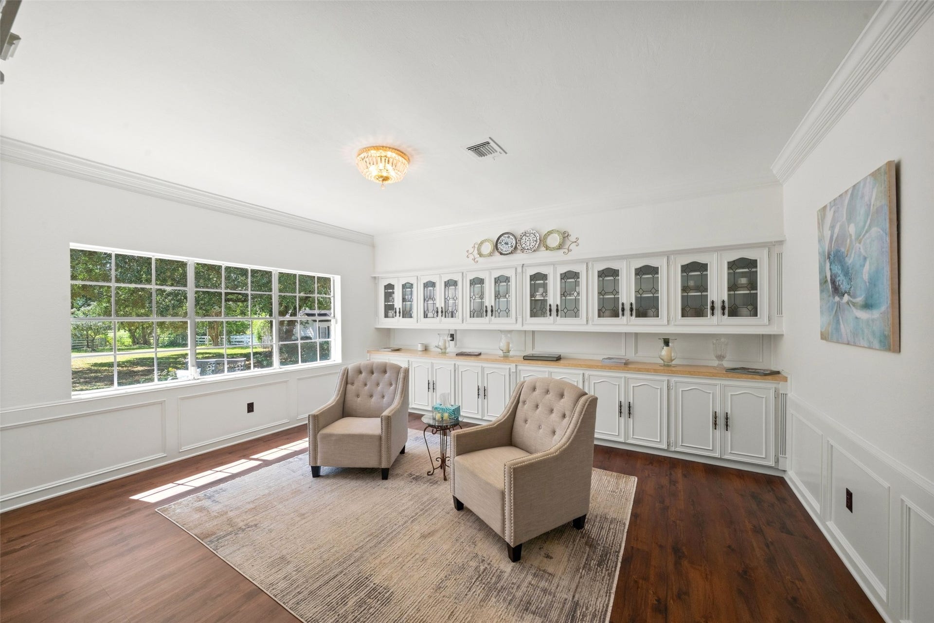 Bright sitting area with a wide bank of windows, wood flooring, and a chandelier overlooking the surrounding greenery.