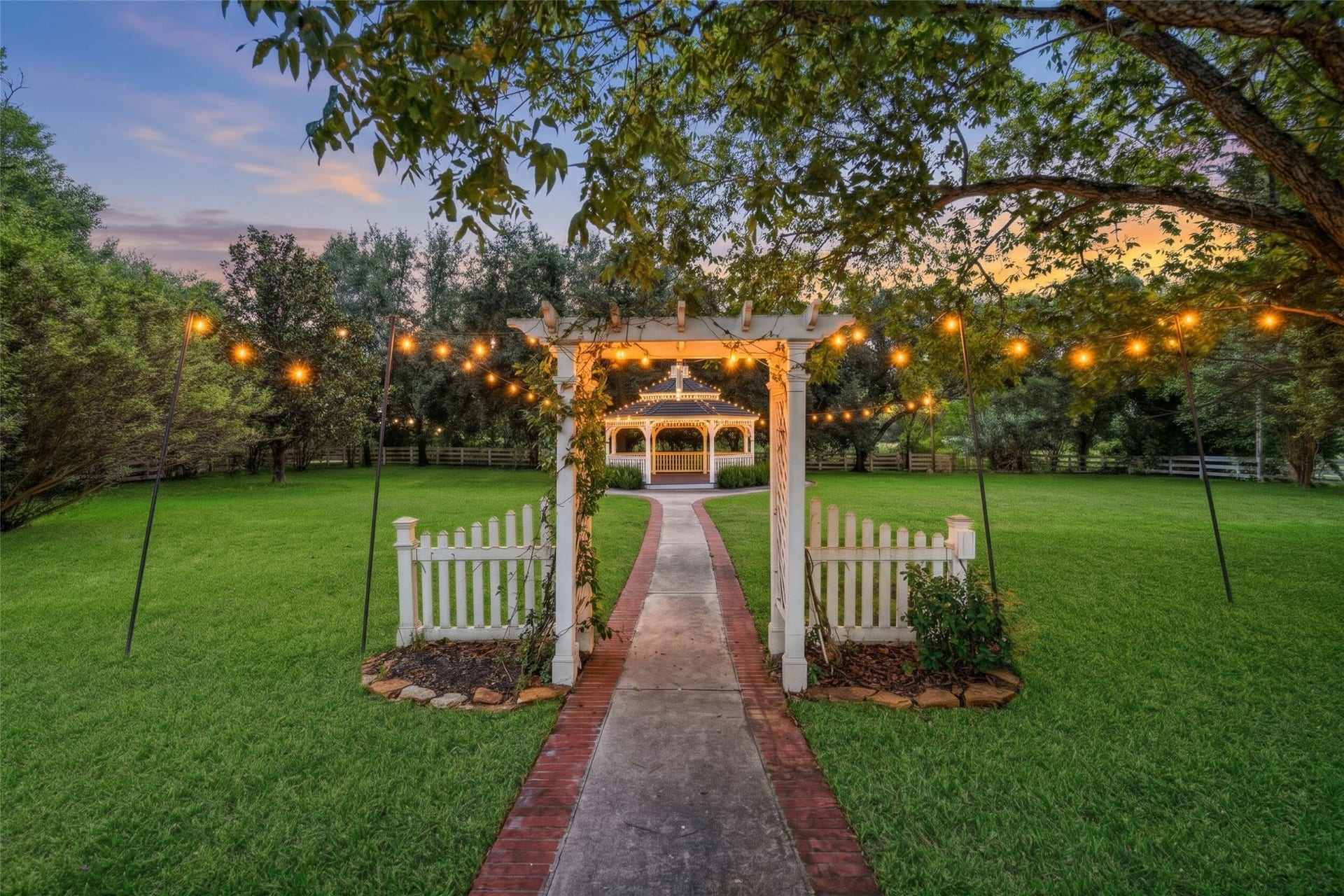 Inviting outdoor ceremony setting with a white gazebo, picket fencing, and overhead string lights beneath a canopy of trees.