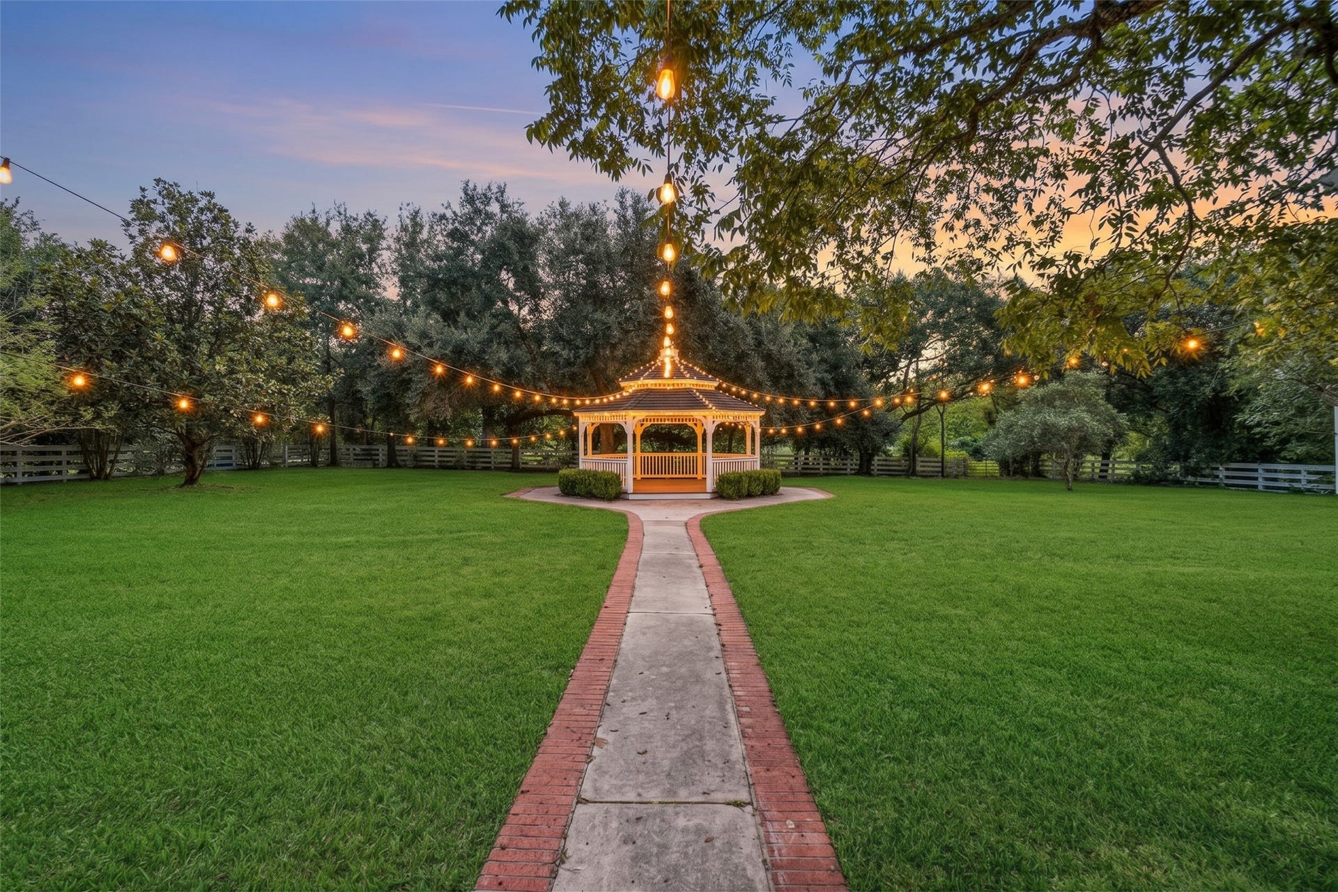 Brick walkway leads to a white garden gazebo, surrounded by open lawn, mature trees, and glowing string lights.