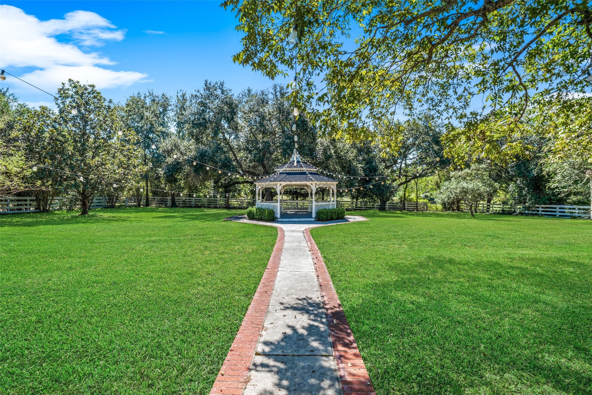 Landscaped walkway leads to a white gazebo, surrounded by expansive lawn and mature trees, creating a scenic setting for outdoor gatherings.