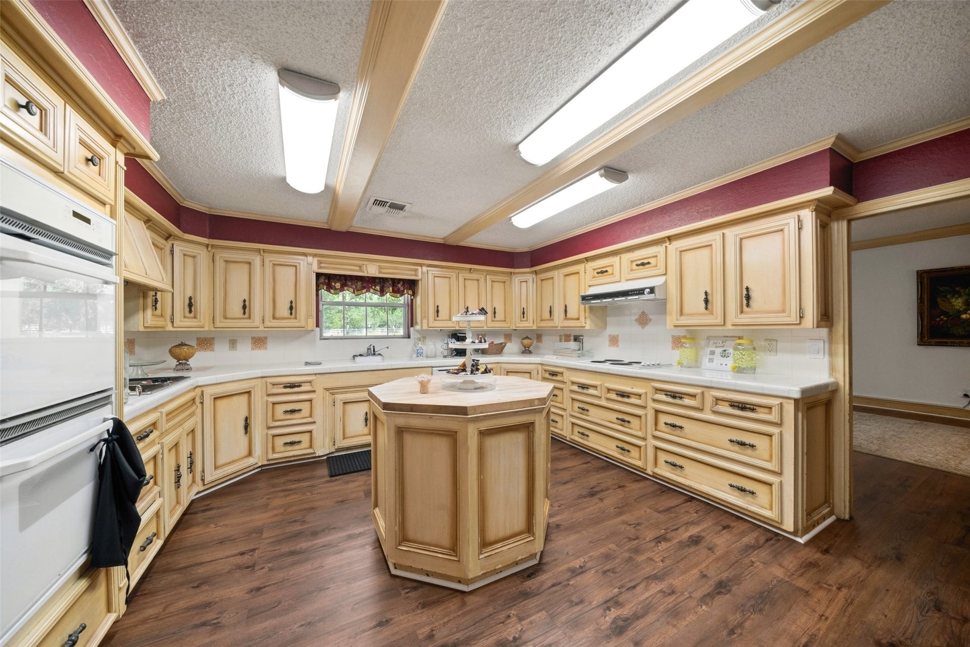 Spacious kitchen features abundant cabinetry, a center island, double ovens, tile countertops, and wood-look flooring, with a pass-through connecting to the adjoining living area.