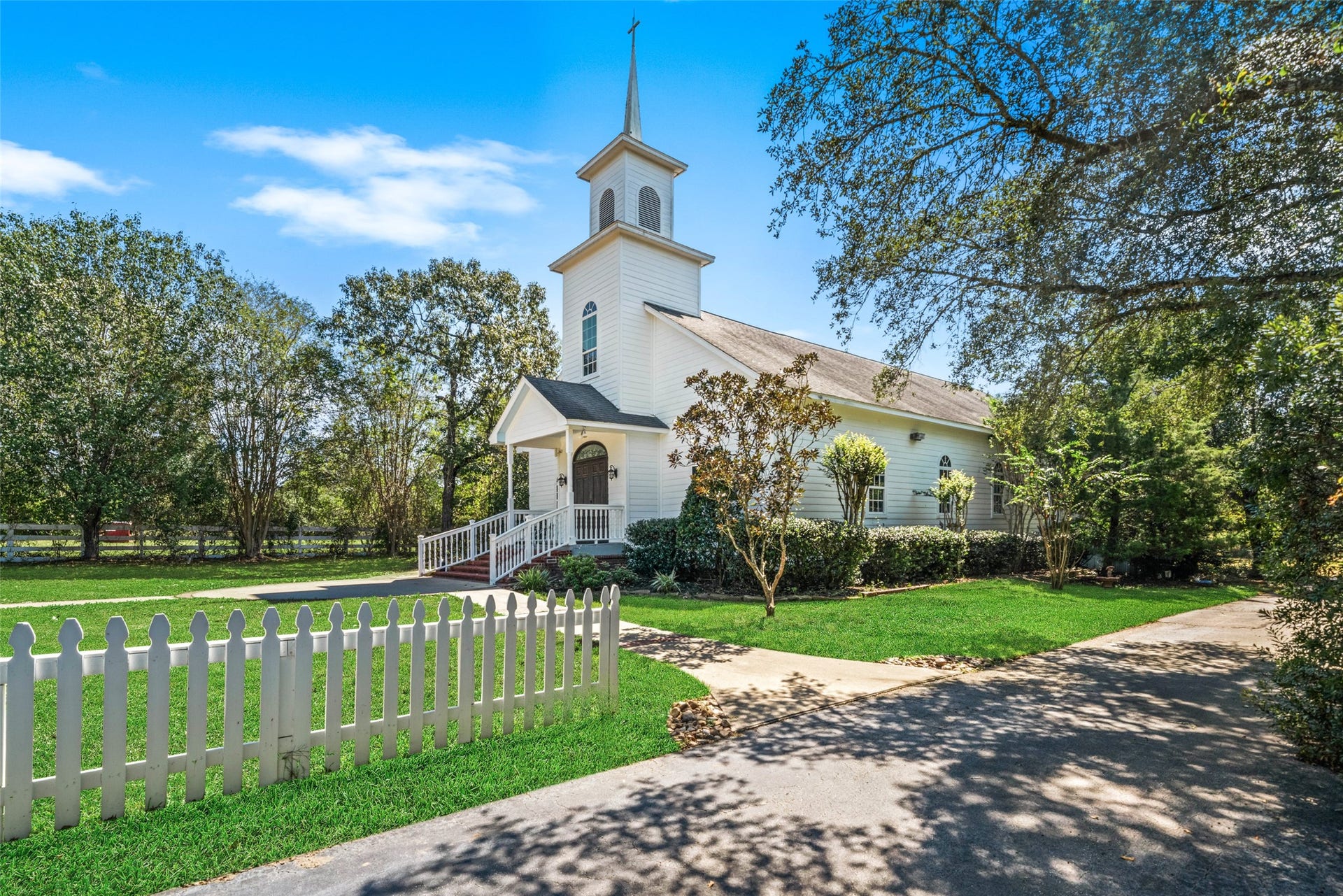 White chapel with a steeple, covered entrance, and picket fencing sits among mature trees and landscaped grounds.