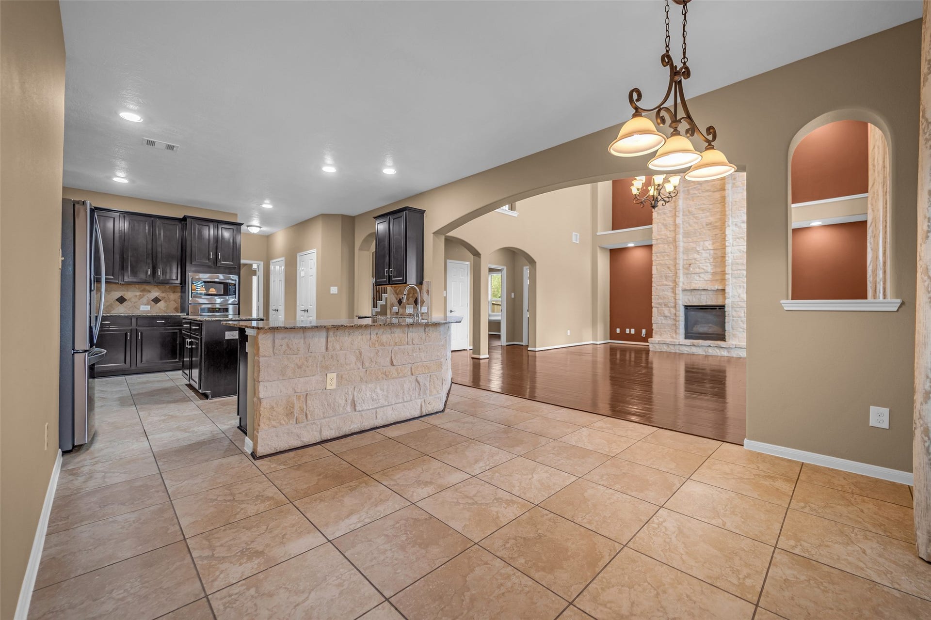 7907 Dunlap Field Lane, Richmond, TX 77407 Spacious open-concept living area featuring a modern kitchen with dark cabinetry, stone accents, and stainless steel appliances, flowing into a cozy living room with a striking stone fireplace and elegant chandelier.
