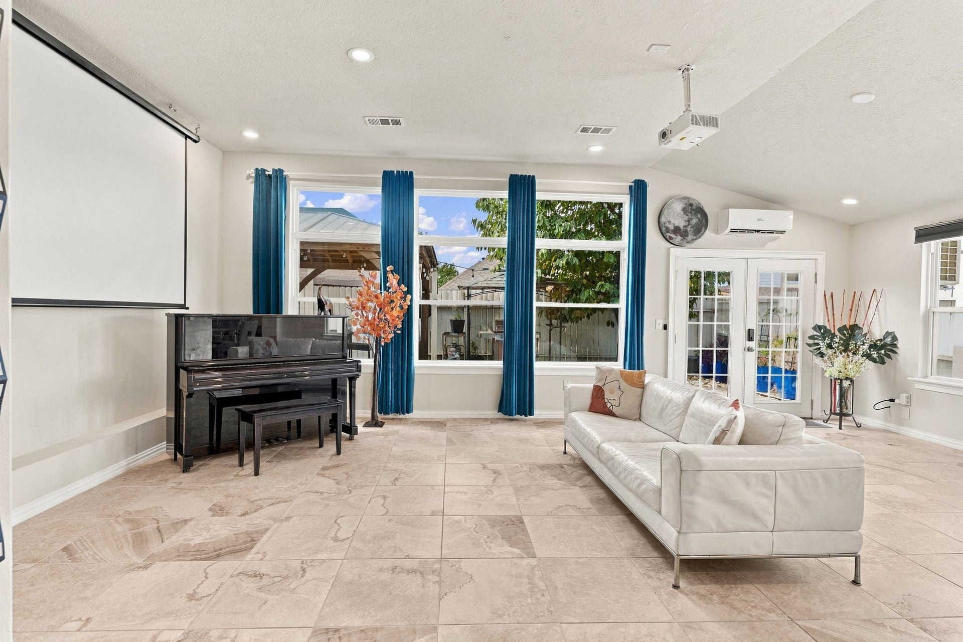Spacious living area with large windows, elegant tile flooring, and a cozy white sofa. Features a piano and a home theater setup, complemented by vibrant blue curtains and French doors leading outside.
