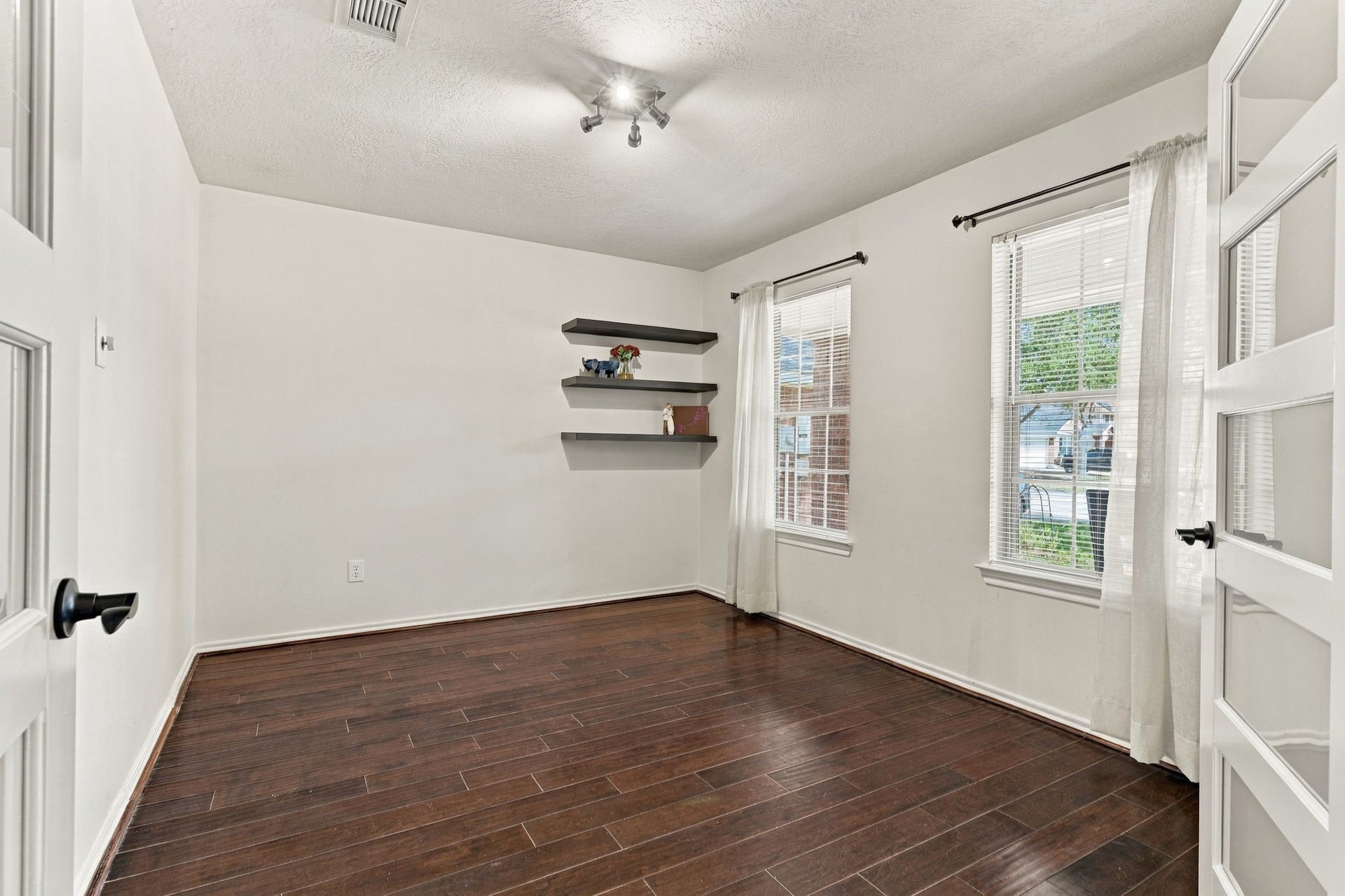 Bright and inviting room featuring rich hardwood floors, ample natural light from large windows, and stylish floating shelves for a modern touch.