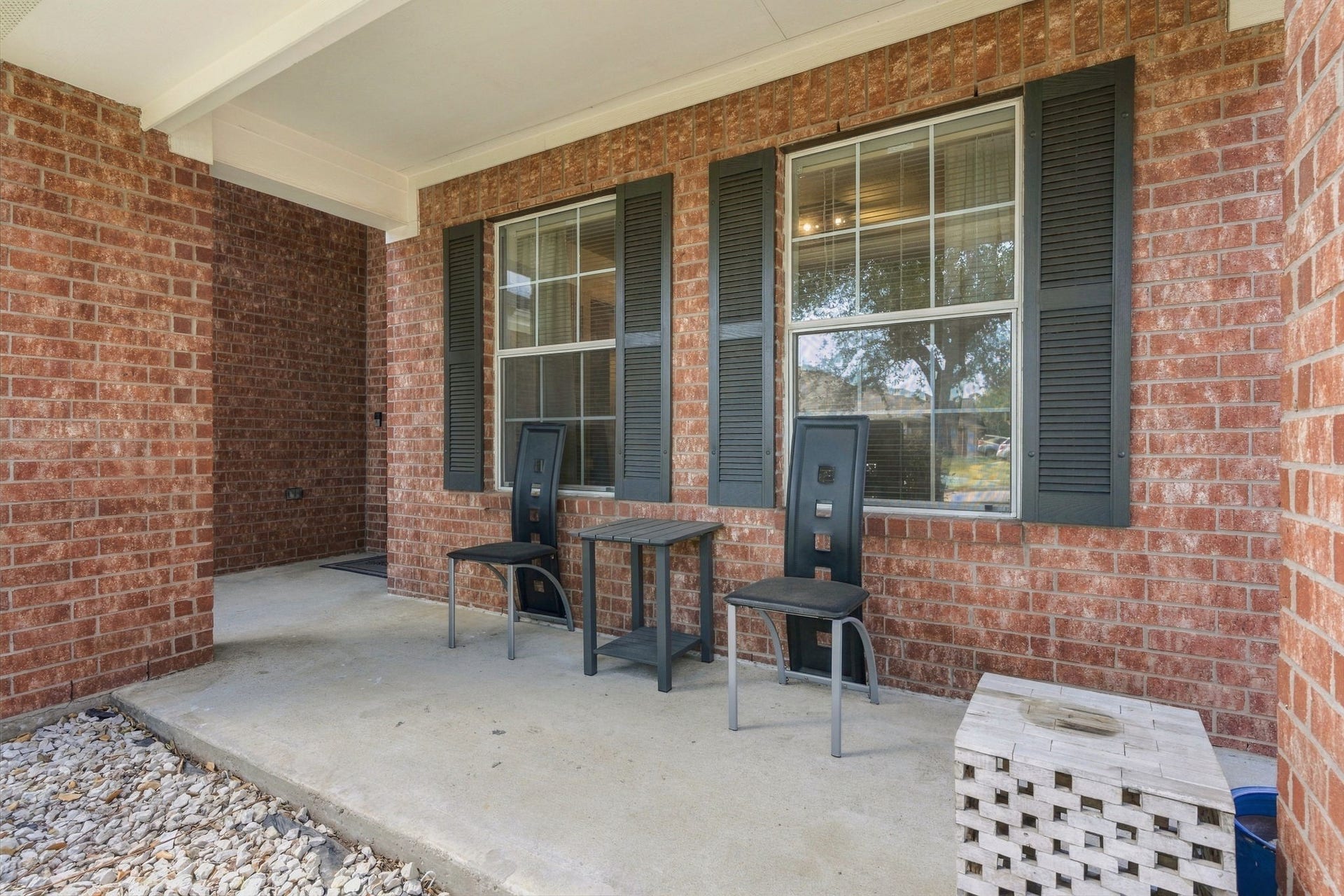 Charming front porch with classic brickwork features cozy seating and large windows with shutters, perfect for relaxing and enjoying the neighborhood view.