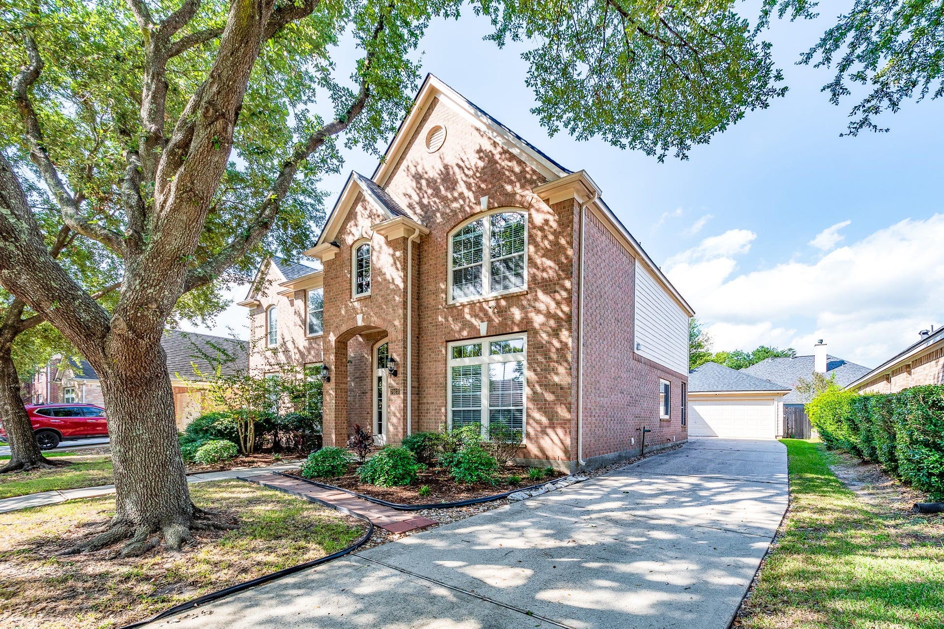 7023 Timber Edge Lane Lane, Humble, TX 77346 Charming two-story brick home with elegant arched windows, nestled under lush trees and featuring a spacious driveway leading to a detached garage.