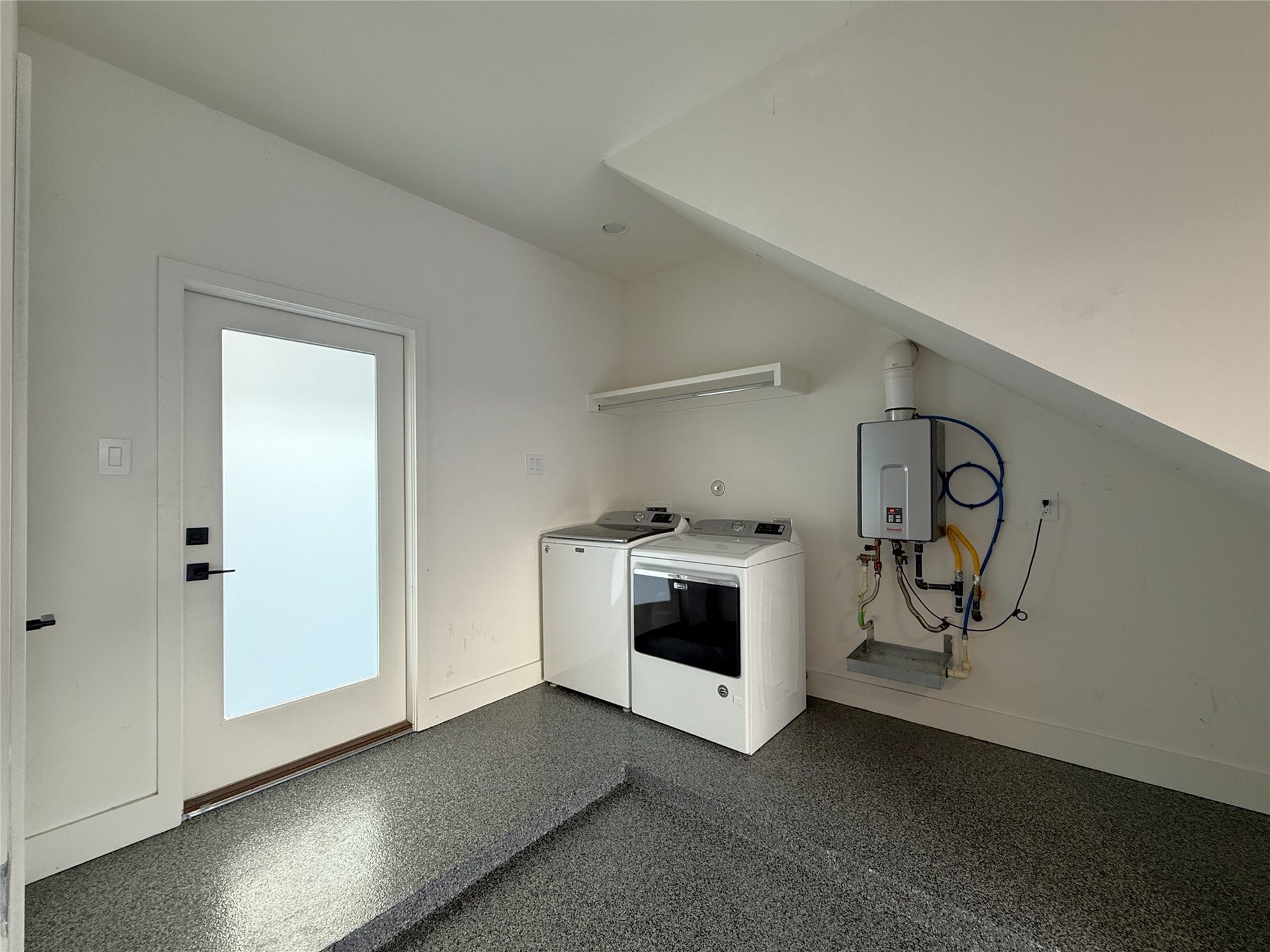 In the garage, the washer/dryer space has an overhead hanging bar. Next to the washer/dryer is the tankless water heater for instant hot water. Beyond the glass paned door is the graveled back yard.