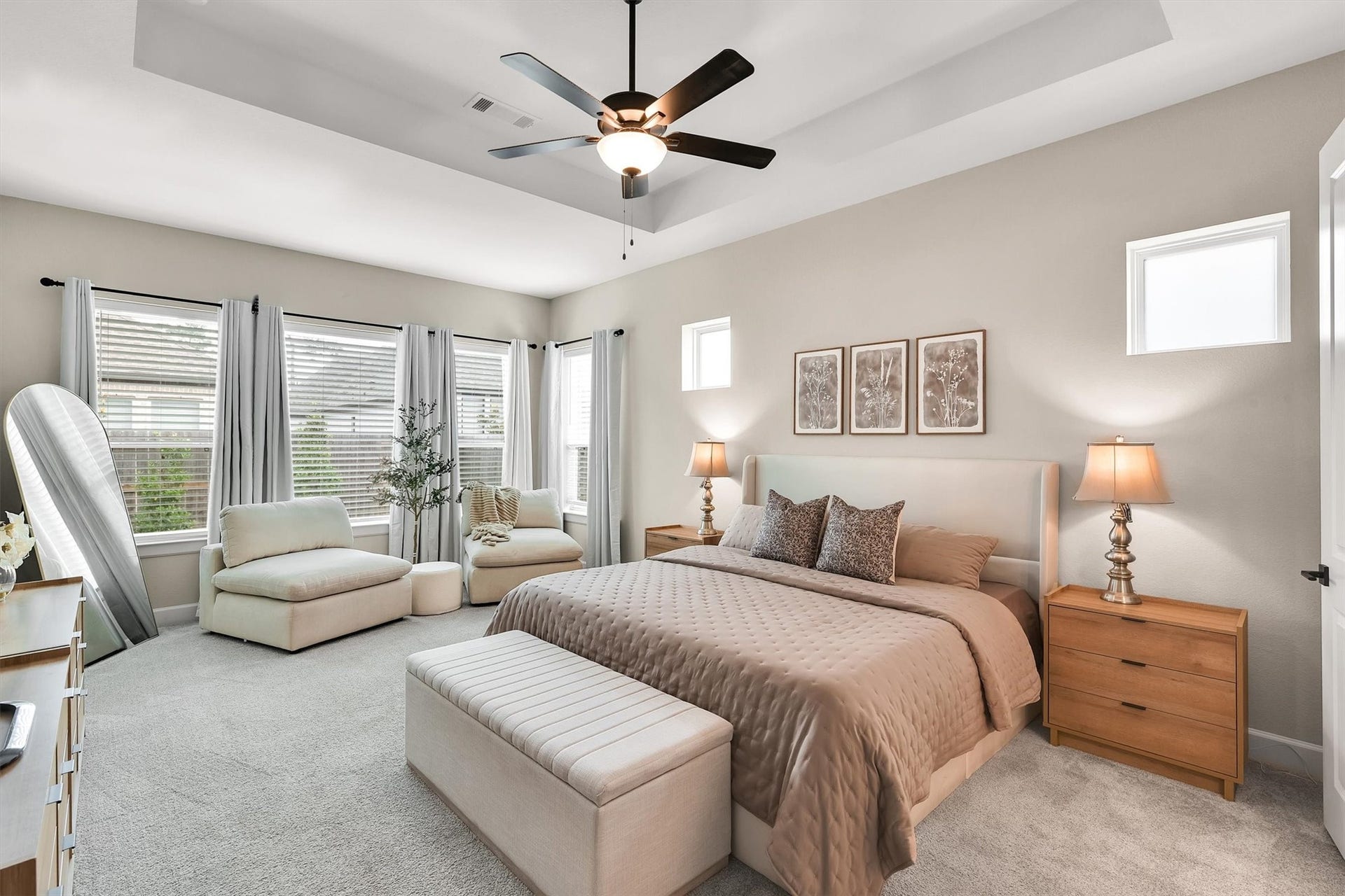 Spacious bedroom features a pastel color scheme, large windows for natural light. Seating area and king sized bed fits perfectly. The ceiling fan and soft carpeting enhance the room's comfort and style.