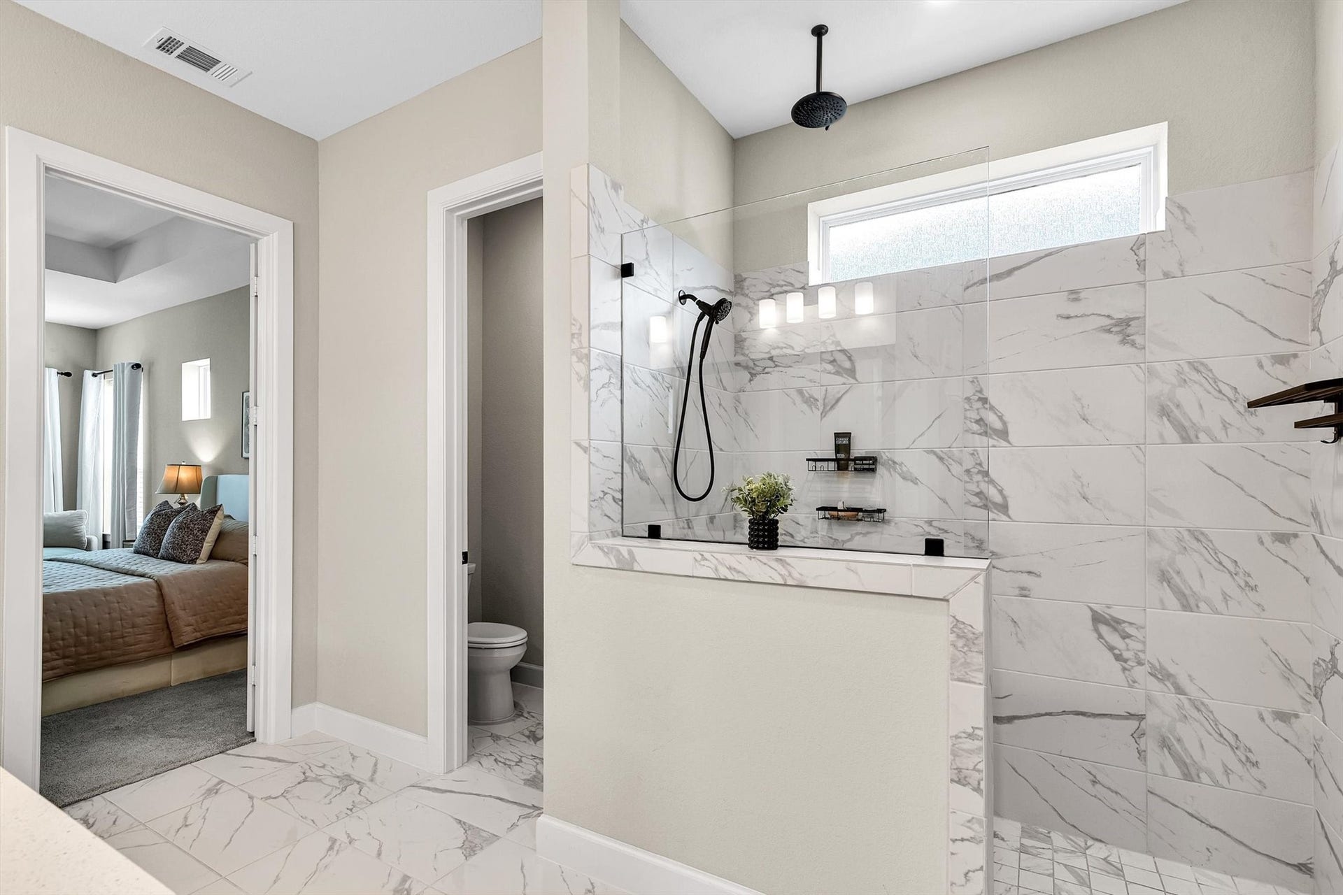 This photo showcases a modern bathroom with a sleek, marble-tiled walk-in shower featuring a rainfall showerhead. Adjacent is the double doorway leading to a cozy, well-lit bedroom with neutral tones and a window.