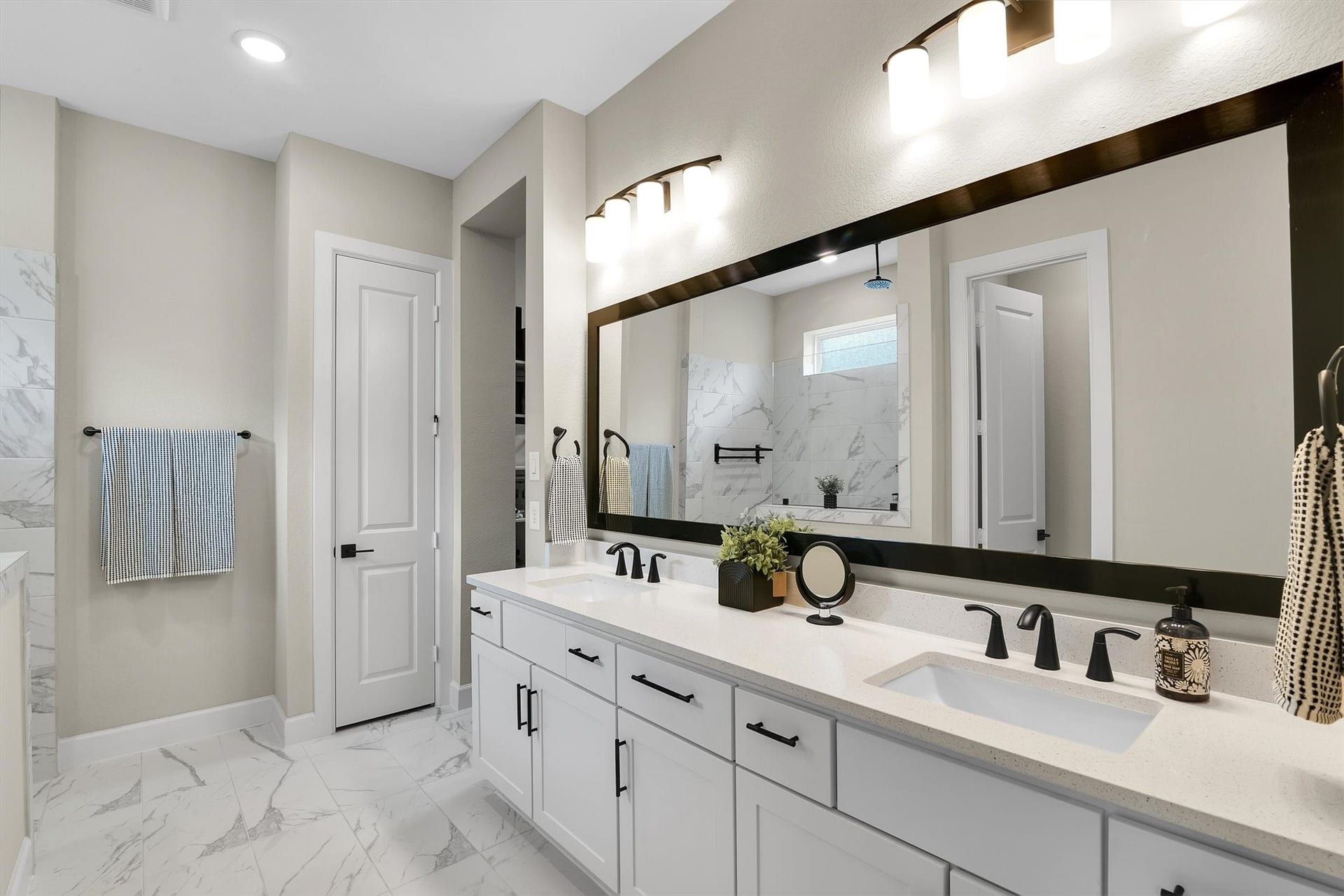 This bathroom features a modern design with a large vanity, dual sinks, and sleek black fixtures. It has a spacious mirror, elegant lighting, and a glass-enclosed shower with marble-style tiles, creating a bright and luxurious atmosphere.