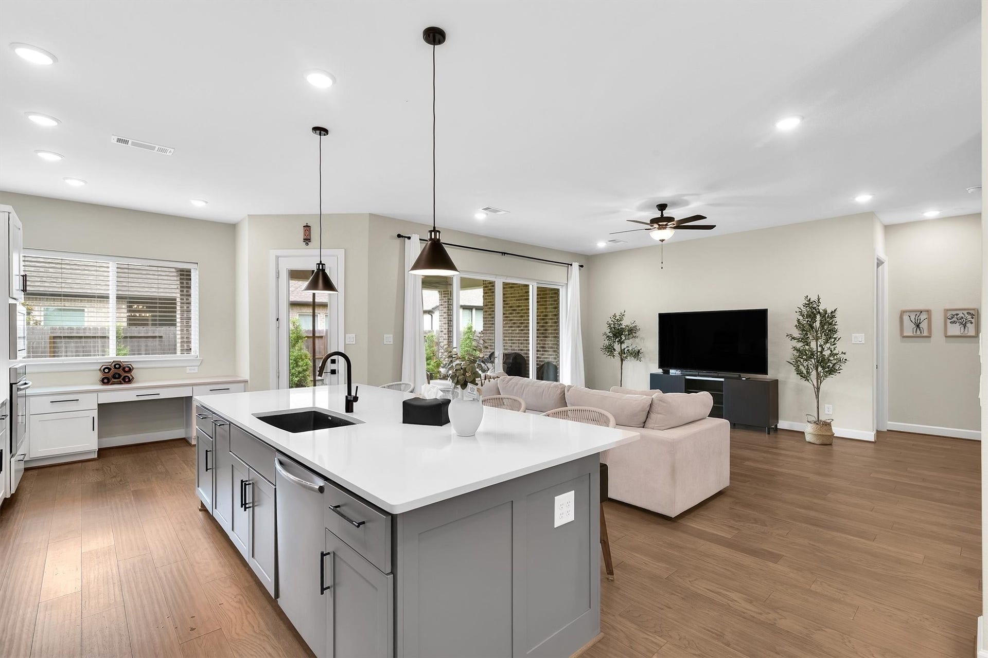 This open-concept living space features a modern kitchen with a large island, sleek cabinetry, and pendant lighting. The adjoining living area includes a cozy seating space, ceiling fan, and a TV setup, all accented by wood flooring and abundant natural light from large windows and a glass door leading to an outdoor area.