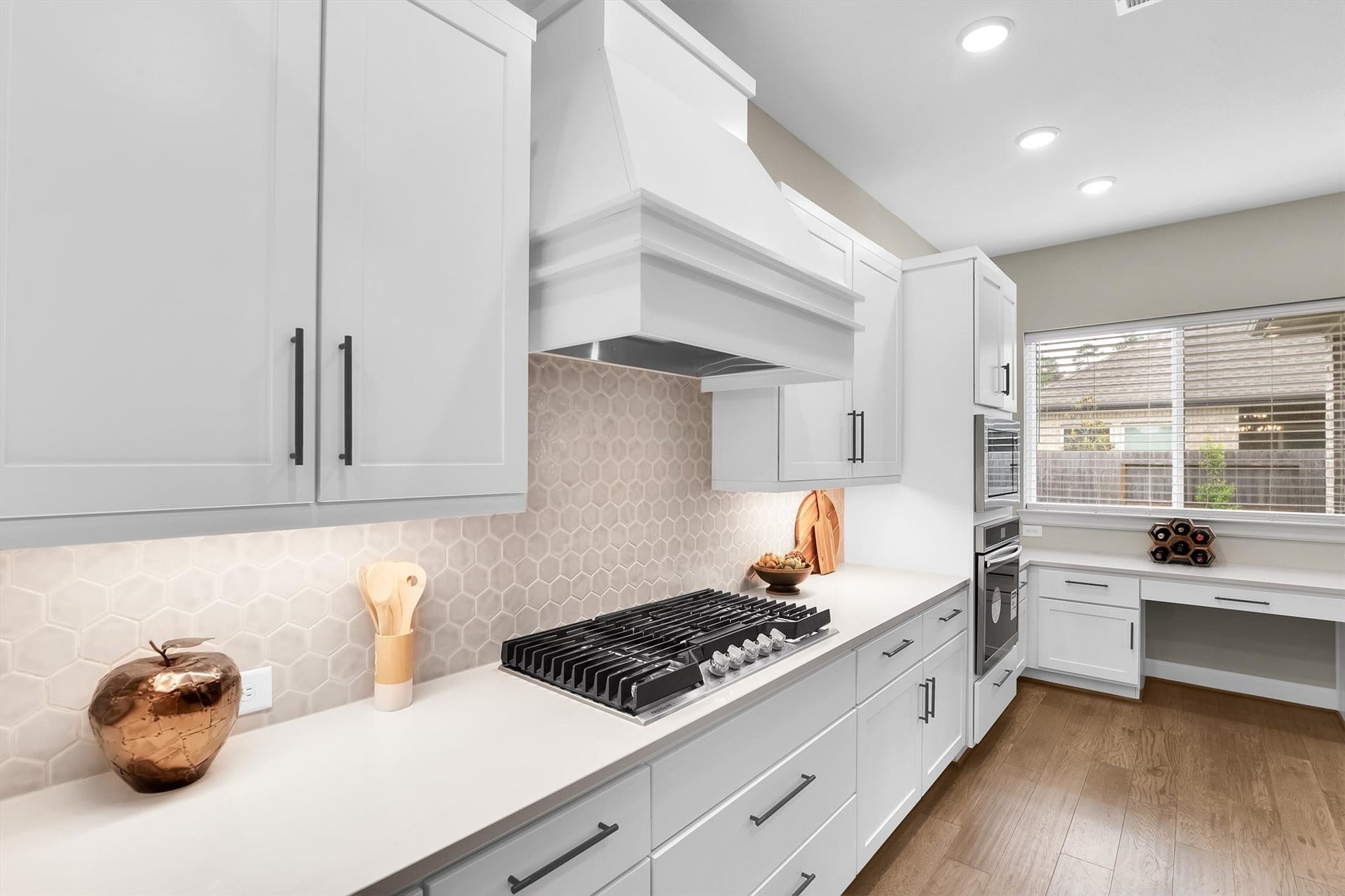 Sleek white cabinetry, hexagonal tile backsplash, and a built-in gas cooktop. It offers ample natural light through a large window, complemented by a spacious countertop and stylish wood flooring.