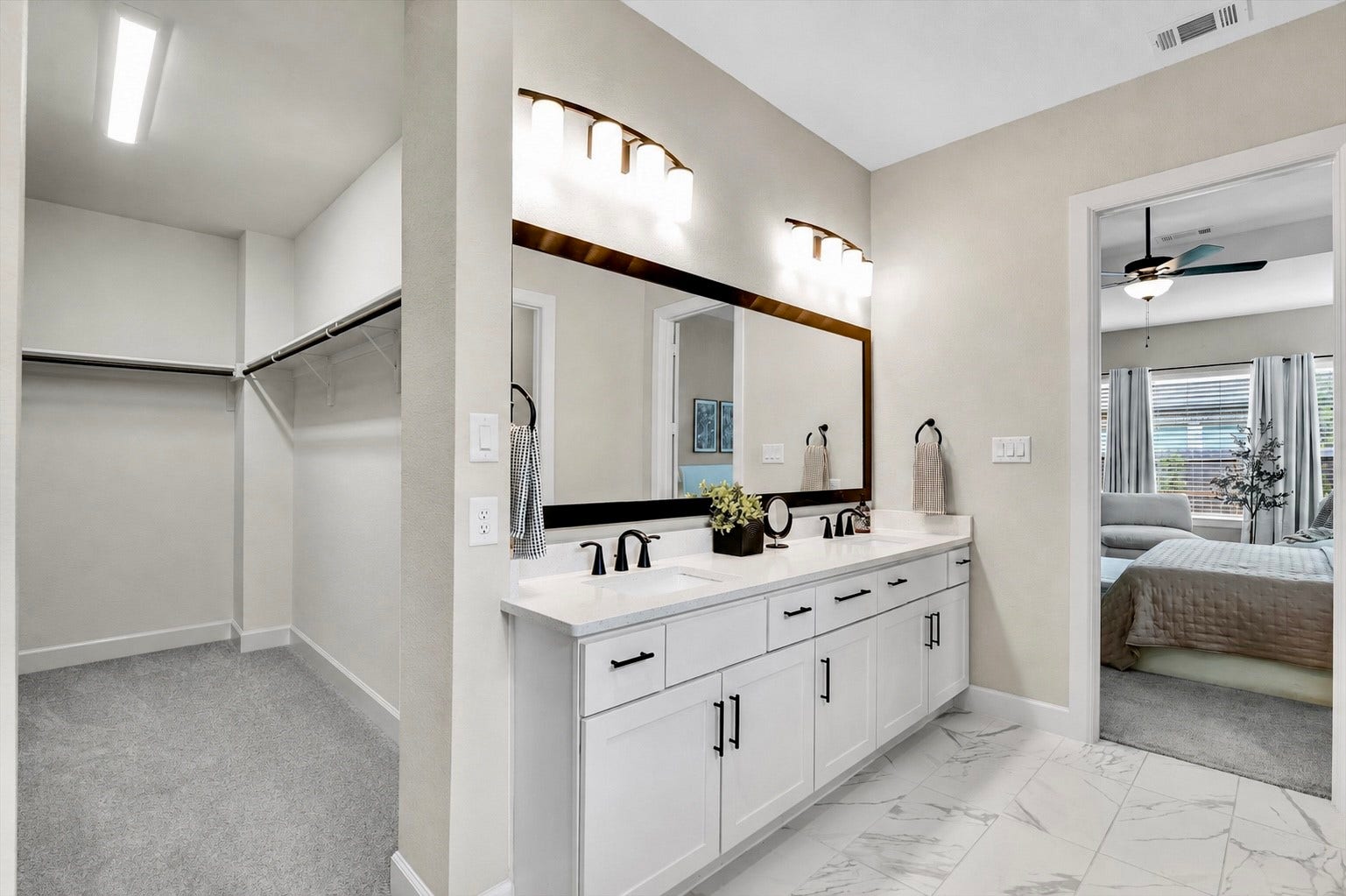 Bright, modern bathroom with dual sinks and sleek lighting opens to a spacious walk-in closet and a cozy bedroom with large windows.