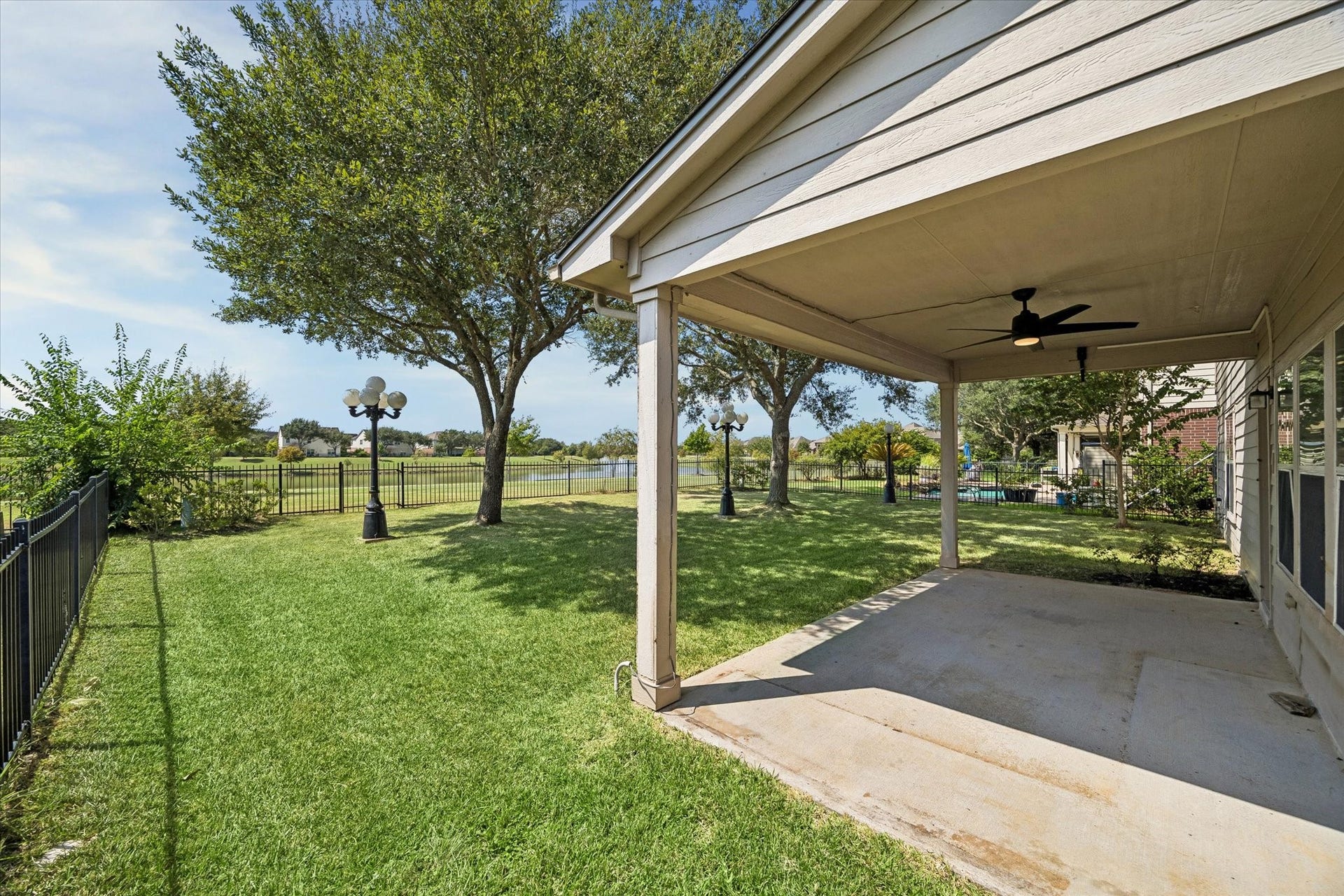 Spacious backyard featuring a covered patio with a ceiling fan, overlooking a manicured lawn and scenic views beyond a charming wrought iron fence.