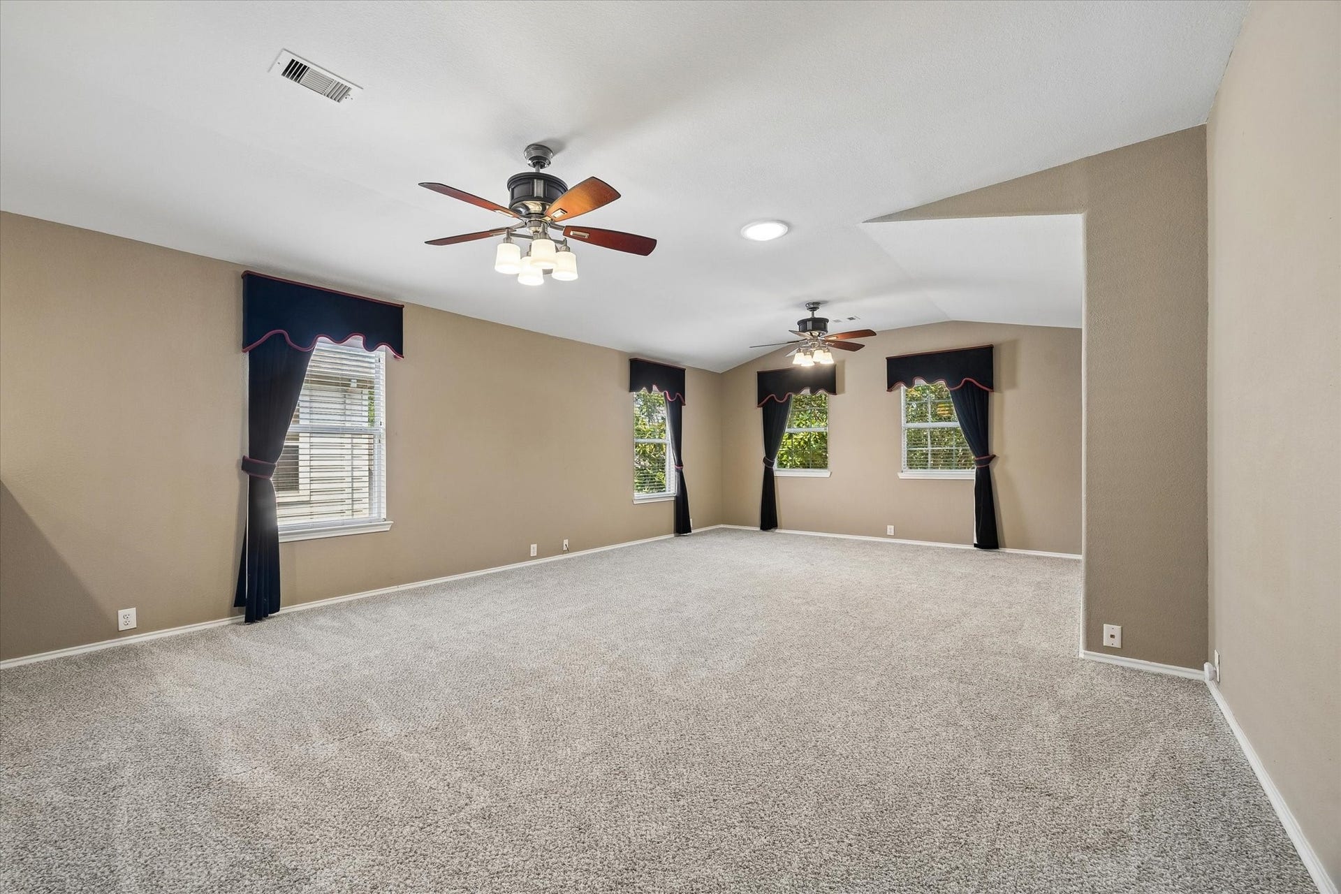 Spacious carpeted game room or living room with neutral walls, featuring large windows and two ceiling fans for optimal comfort.