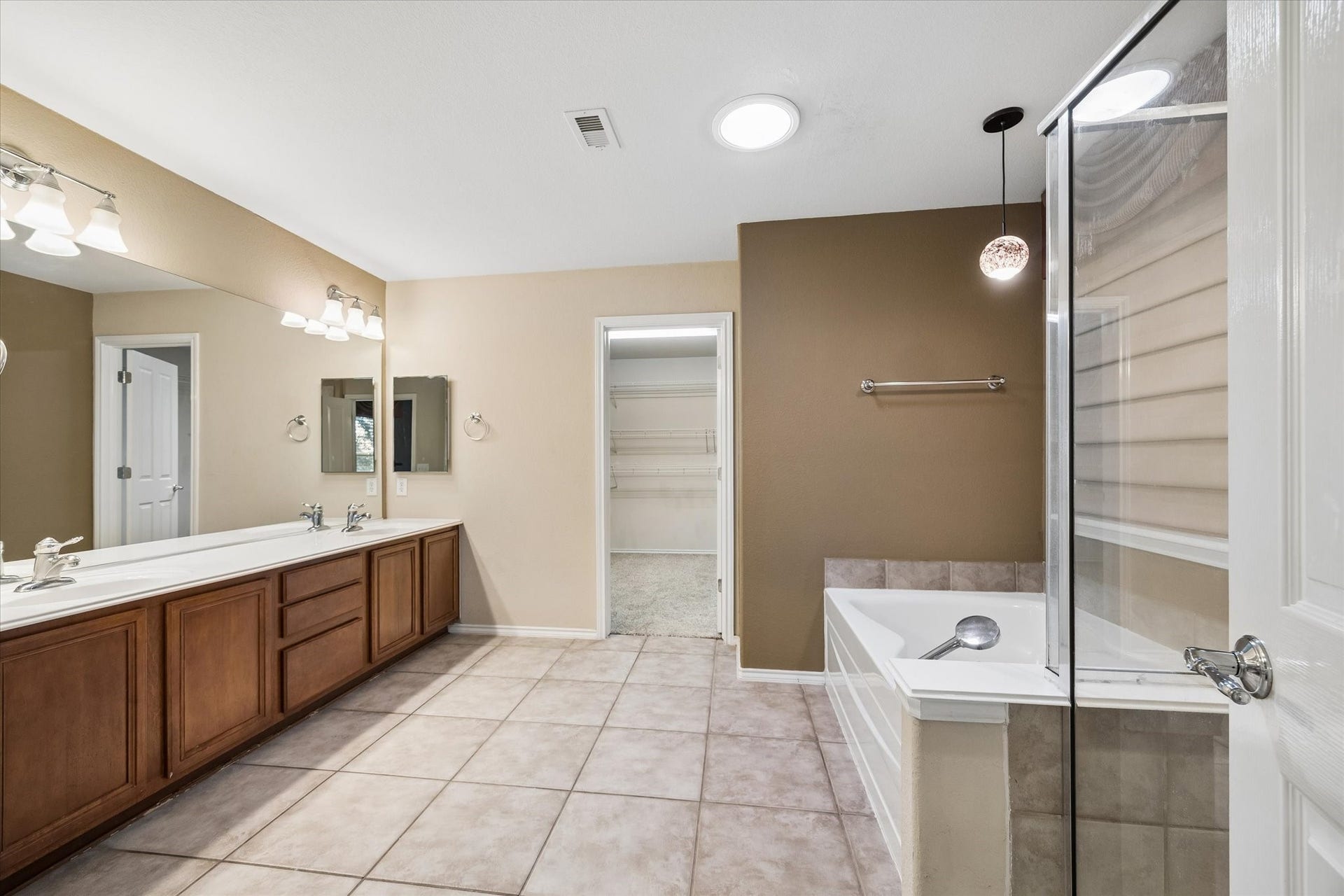Spacious primary bathroom with dual sinks, elegant wooden cabinetry, a large soaking tub with a separate glass-enclosed shower, and direct access to a walk-in closet.