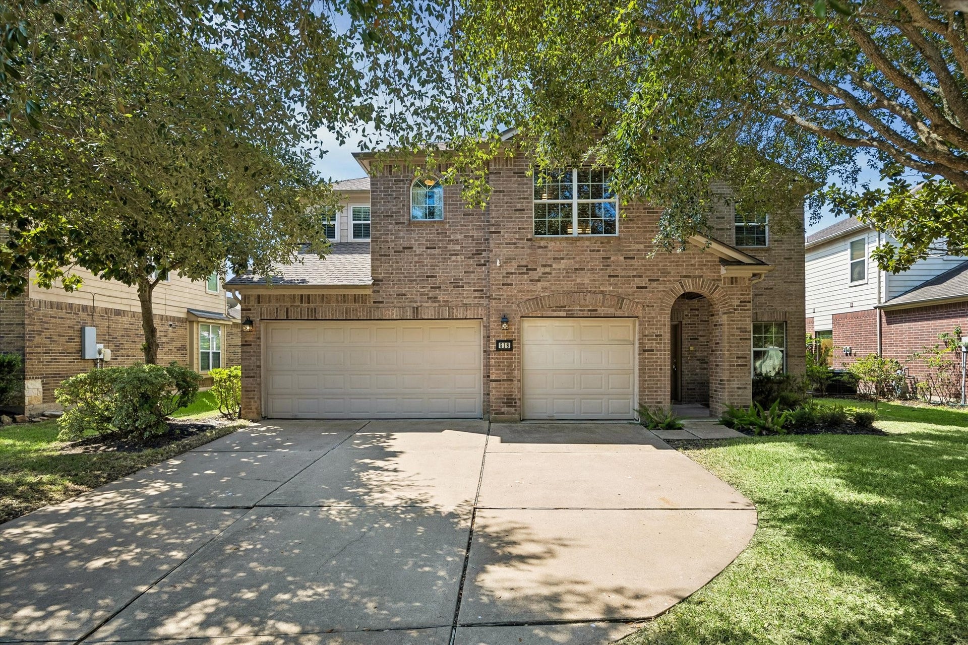 Charming two-story brick home with a spacious three-car garage, nestled among mature trees, offering ample driveway space and lush green landscaping.
