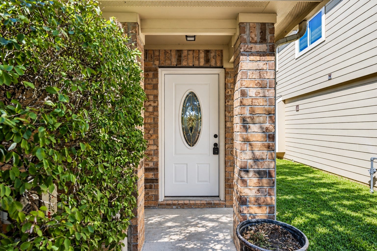 Welcoming front entryway featuring a classic brick column, mature landscaping, and a secure front door with a decorative glass insert.